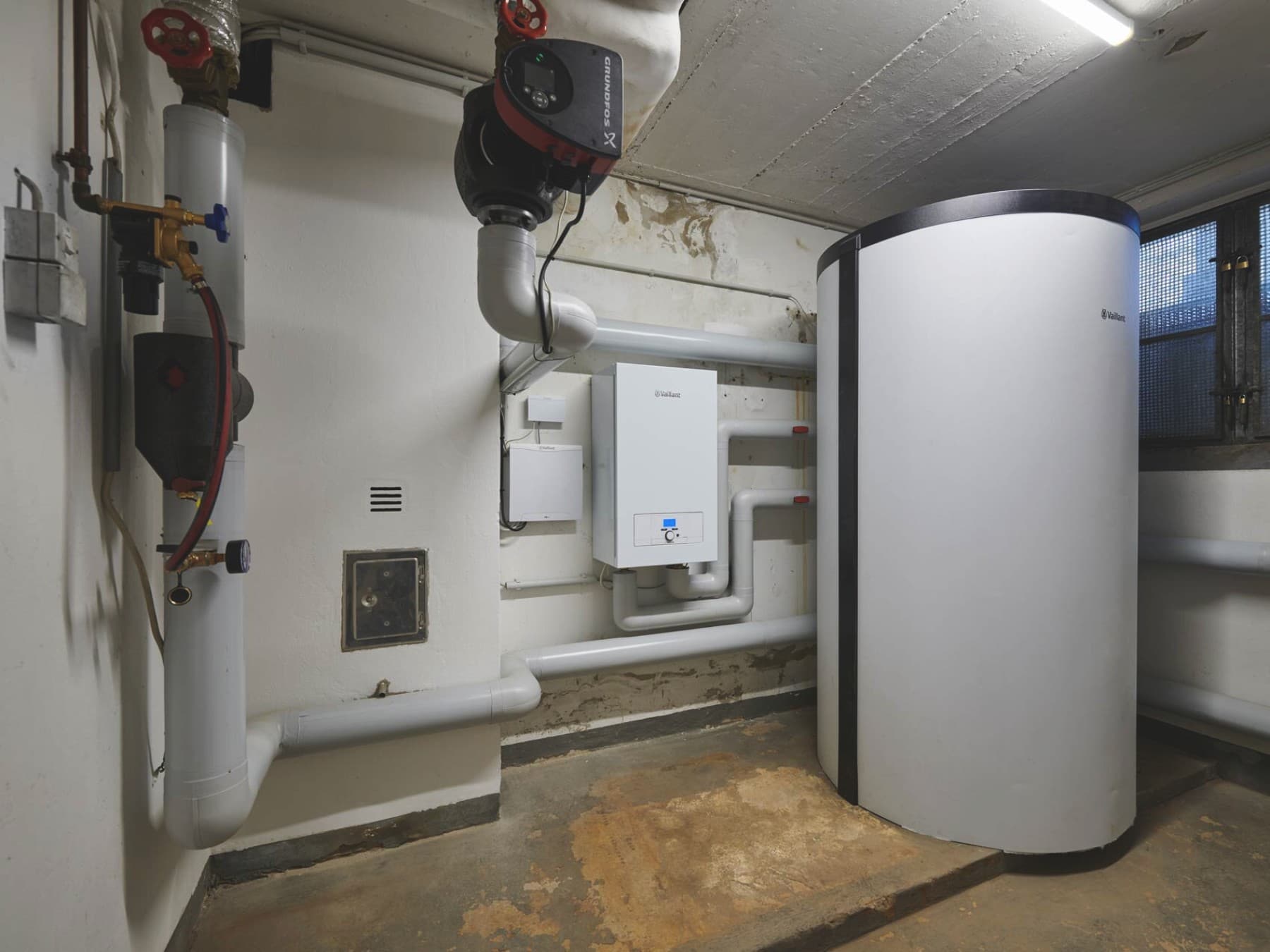 Heizungsraum im Keller mit an der Wand montierten Rohrleitungen. Ein großer 800 Liter Pufferspeicher steht auf der rechten Seite und links daneben ein Elektrozusatzheizgerät an der Wand vom Typ eloBLOCK von Vaillant.