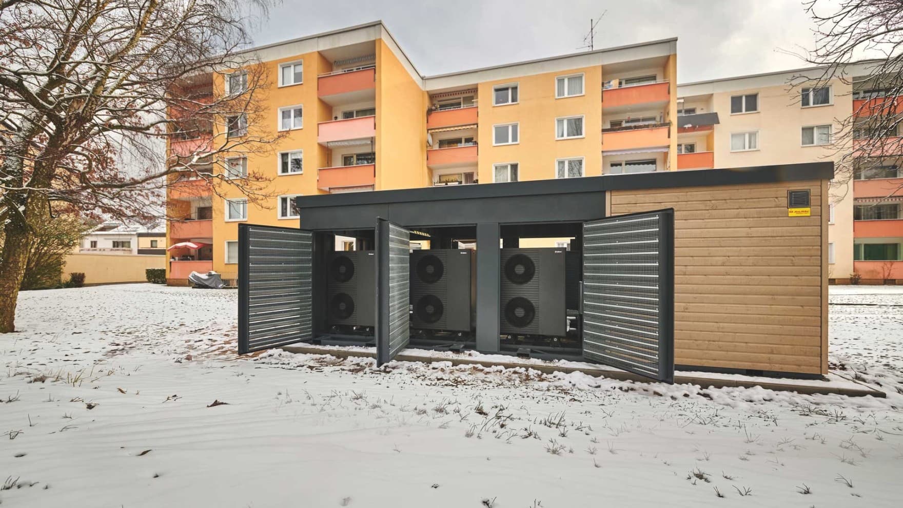 Außen aufgestellte kompakte Heizzentrale Energy Unit steht mit geöffneten Türen und mehreren Luft/Wasser-Wärmepumpen von Vaillant vor zwei Mehrfamilienhäusern auf einem Betonfundament. Sie ist umgeben von verschneiter Grünfläche.