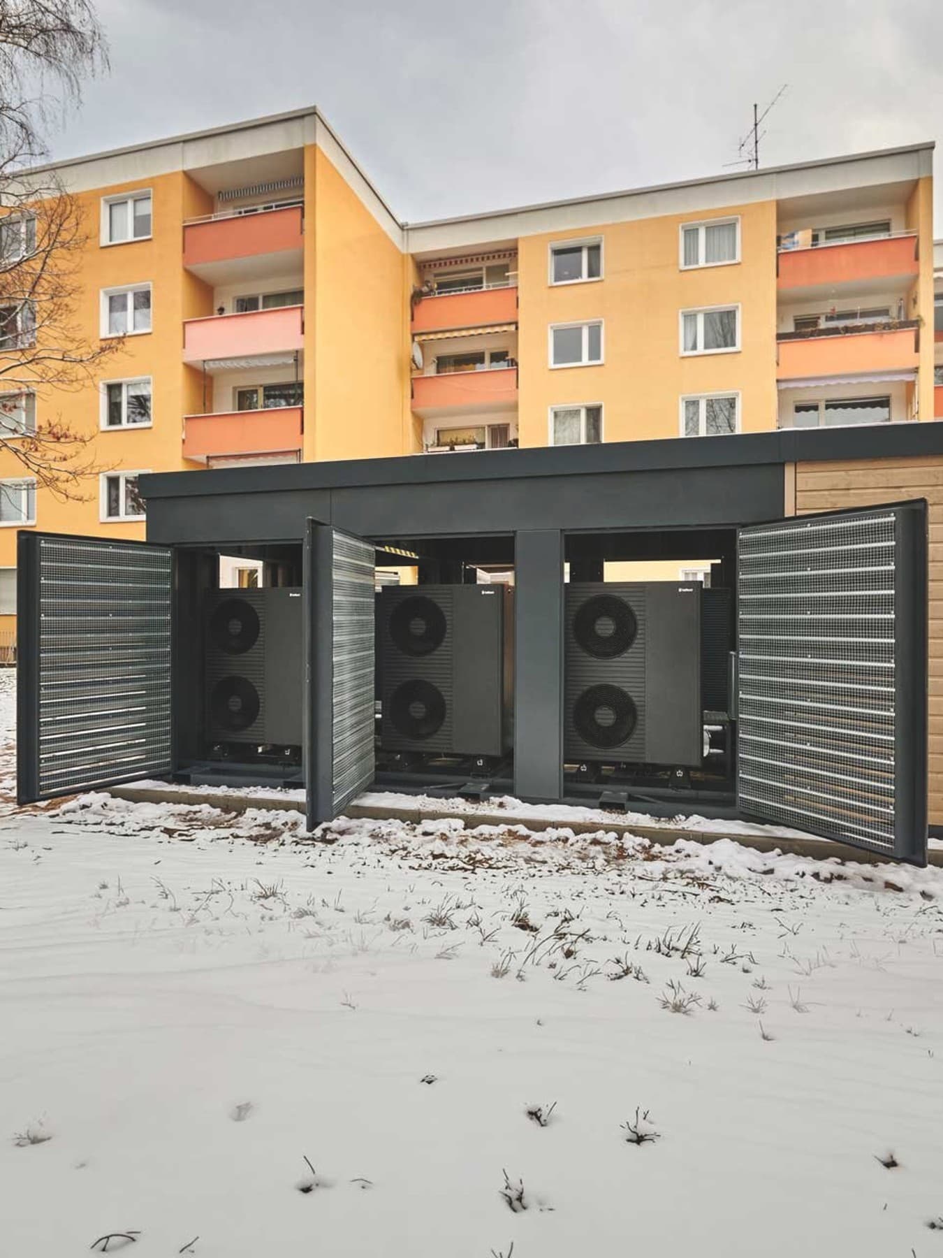 Außen aufgestellte kompakte Heizzentrale Energy Unit steht mit geöffneten Türen und mehreren Luft/Wasser-Wärmepumpen von Vaillant vor zwei Mehrfamilienhäusern auf einem Betonfundament. Sie ist umgeben von verschneiter Grünfläche.