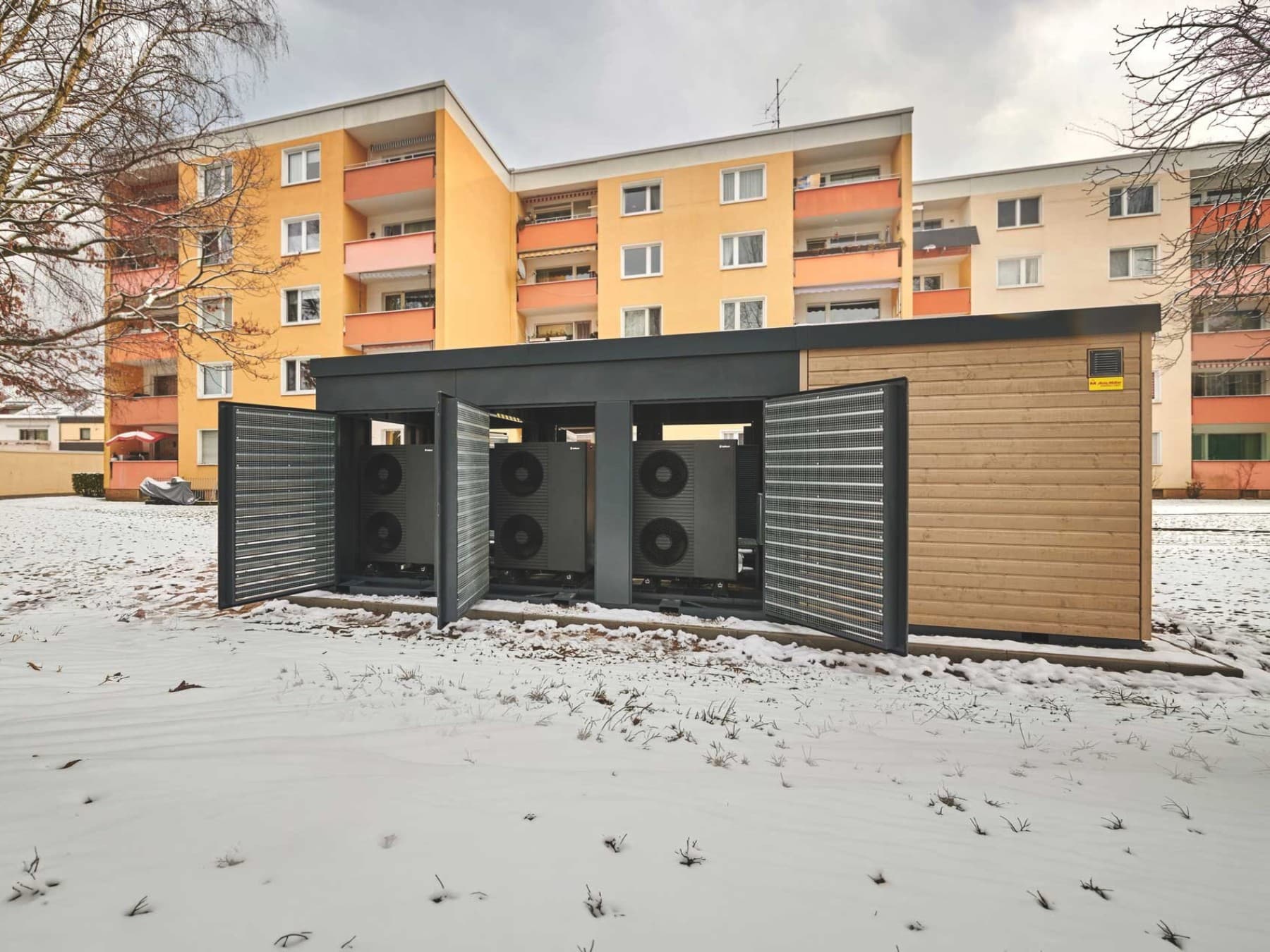 Außen aufgestellte kompakte Heizzentrale Energy Unit steht mit geöffneten Türen und mehreren Luft-Wasser-Wärmepumpen von Vaillant vor zwei Mehrfamilienhäusern auf einem Betonfundament. Sie ist umgeben von verschneiter Grünfläche.