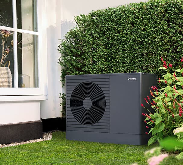 Die neue Vaillant Wärmepumpe aroTHERM pro steht vor einer Hecke auf dem Rasen neben einem Haus und einem hohen Fenster.