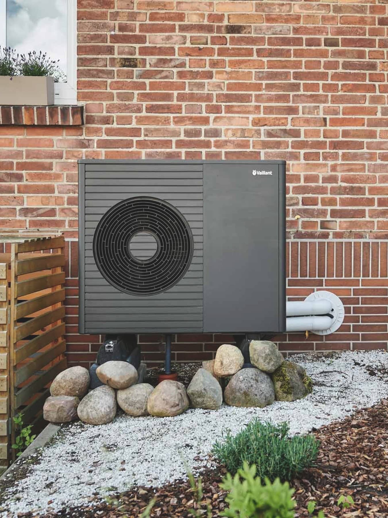 Außeneinheit der Luft-Wasser-Wärmepumpe aroTHERM plus VWL 75/8.1 A von Vaillant vor der Backsteinfassade eines Altbaus mit zwei Fenstern und bepflanztem Vorgarten.