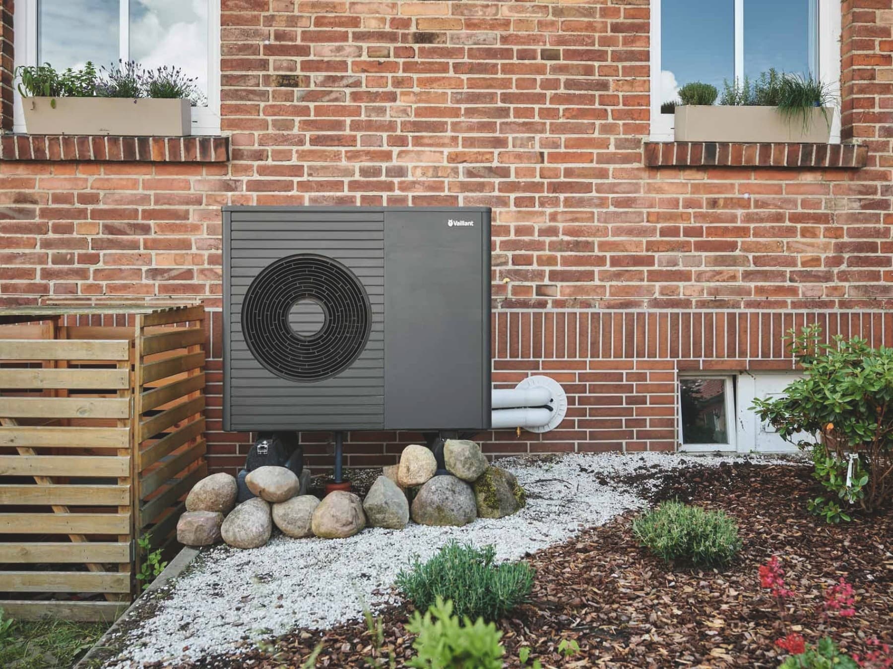 Außeneinheit der Luft-Wasser-Wärmepumpe aroTHERM plus VWL 75/8.1 A von Vaillant vor der Backsteinfassade eines Altbaus mit zwei Fenstern und bepflanztem Vorgarten.