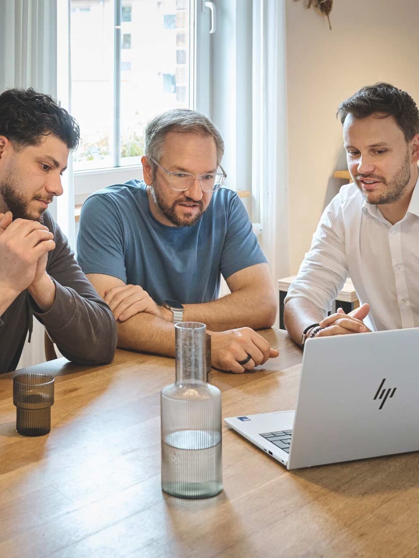 Drei Männer sitzen an einem Holztisch in einer hellen Küche und schauen gemeinsam auf einen geöffneten Laptop. Auf dem Tisch stehen mehrere Gläser, eine Karaffe und eine Tasse.