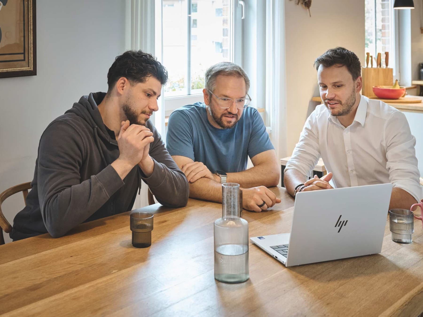 Drei Männer sitzen an einem Holztisch in einer hellen Küche und schauen gemeinsam auf einen geöffneten Laptop. Auf dem Tisch stehen mehrere Gläser, eine Karaffe und eine Tasse.