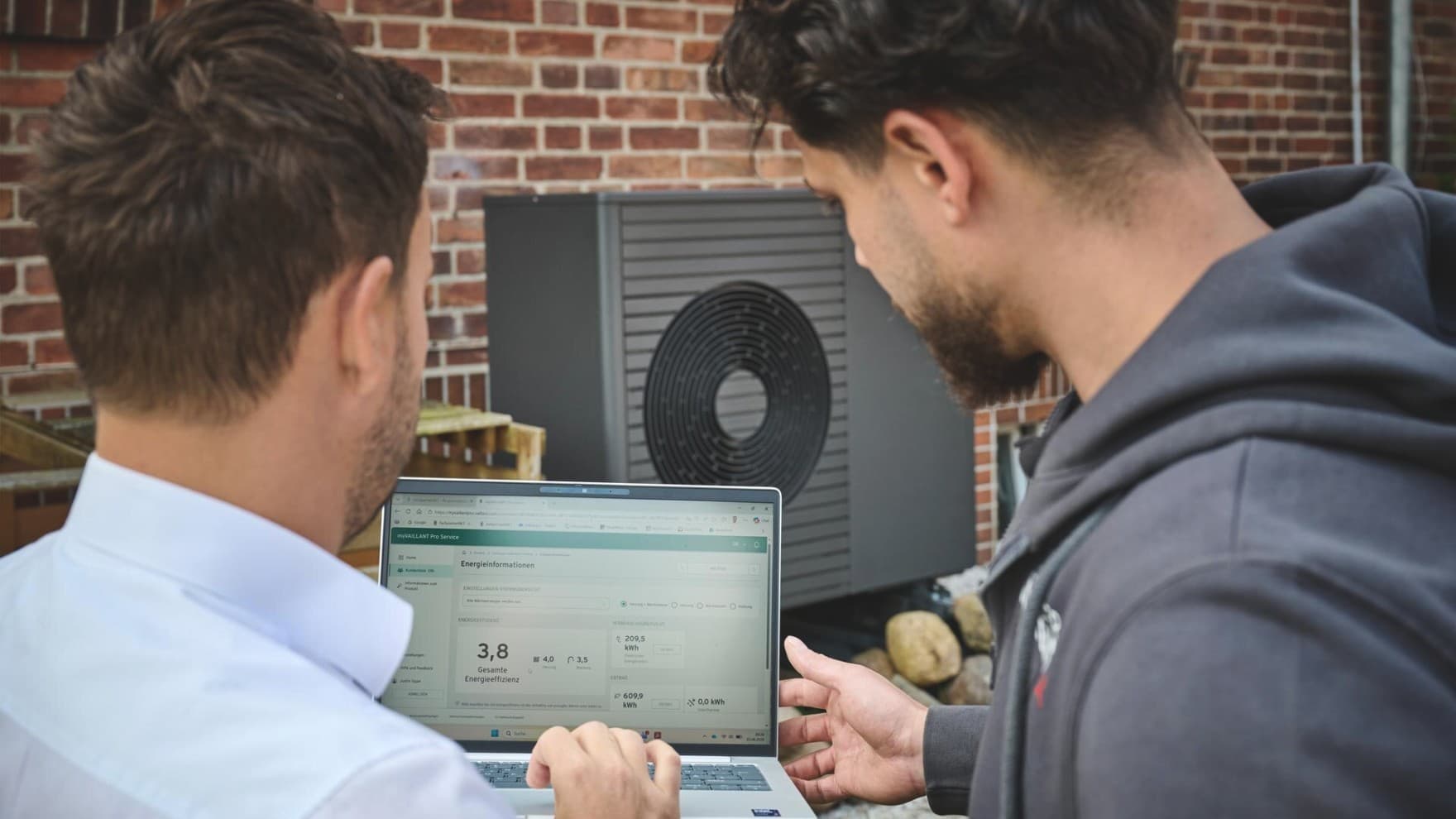 Zwei Personen betrachten auf einem Laptop die Energiedaten einer Luft-Wasser-Wärmepumpe aroTHERM plus VWL 75/8.1 A von Vaillant vor der Backsteinfassade eines Altbaus.