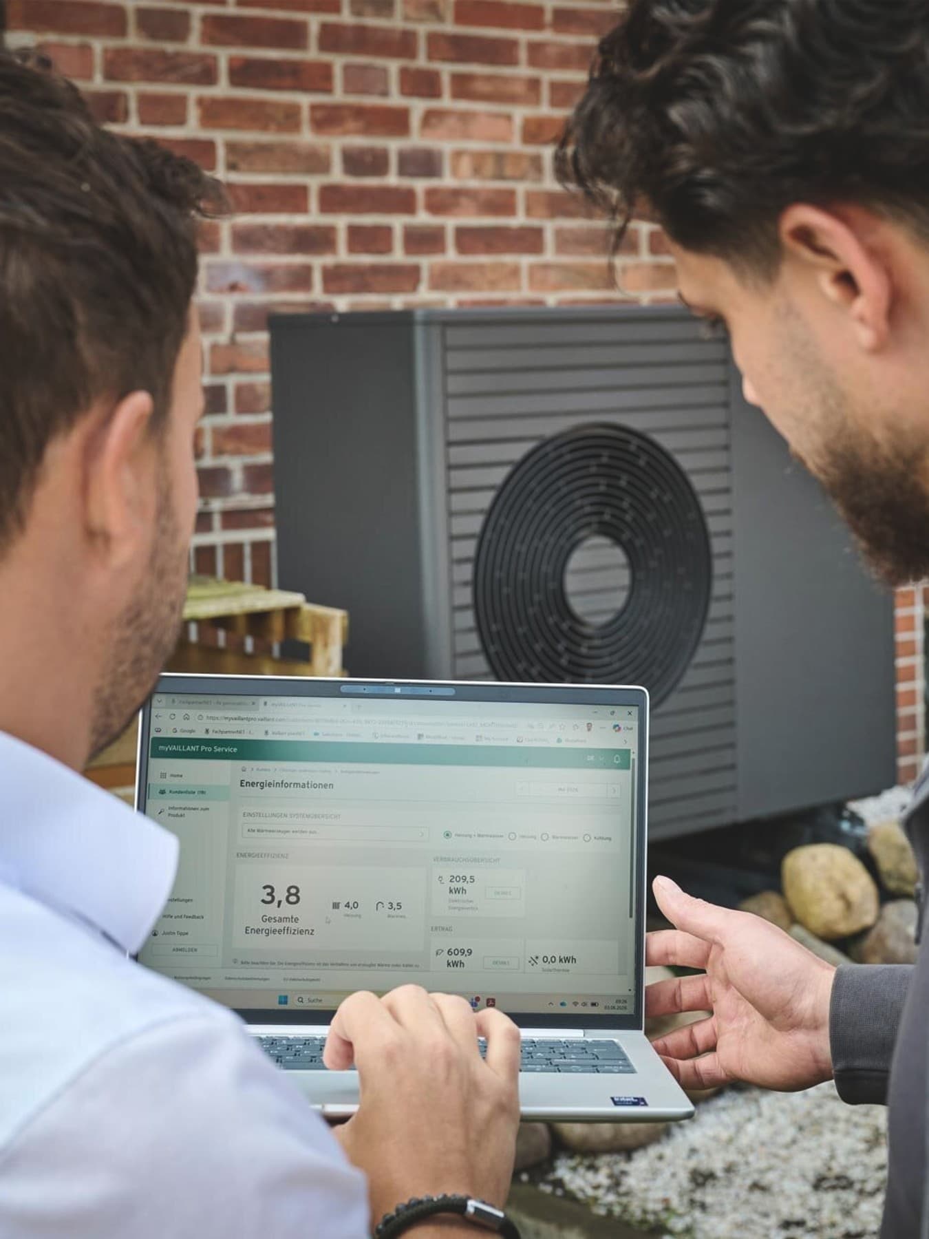 Zwei Personen betrachten auf einem Laptop die Energiedaten einer Luft-Wasser-Wärmepumpe aroTHERM plus VWL 75/8.1 A von Vaillant vor der Backsteinfassade eines Altbaus.