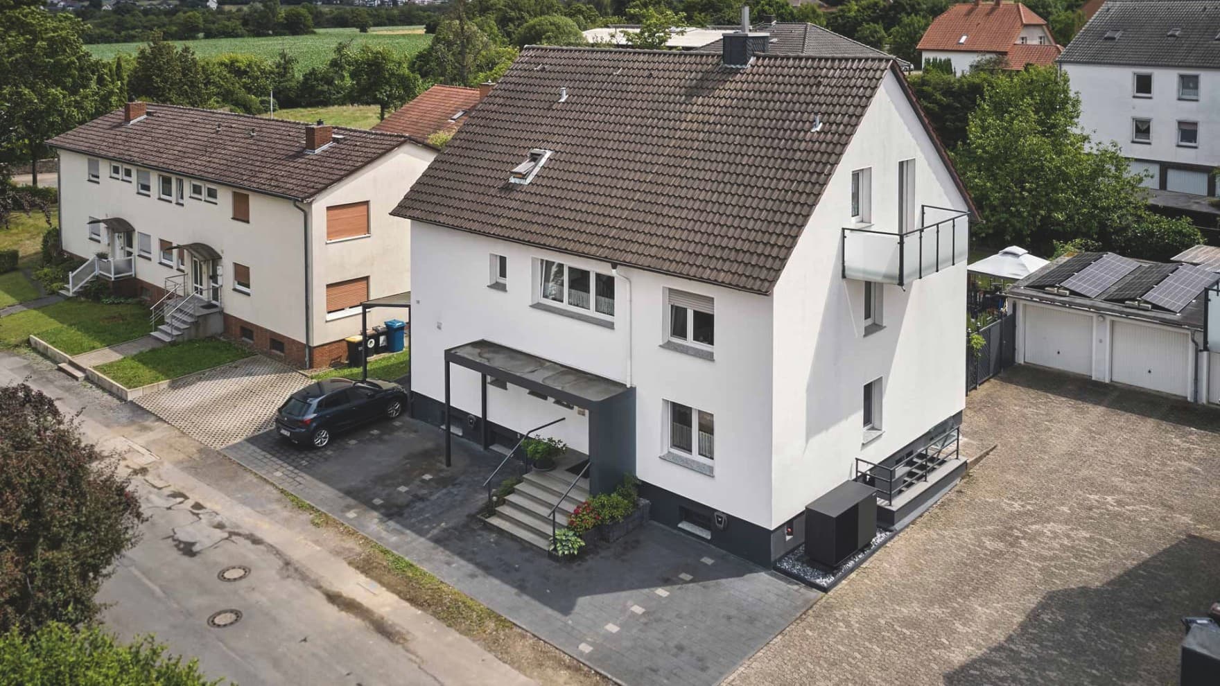 Luftaufnahme eines modernisierten weißen Mehrfamilienhauses mit dunklem Ziegeldach, Balkon und überdachtem Eingangsbereich. Rechts neben dem Gebäude ist die Außeneinheit der Wärmepumpe aroTHERM perform von Vaillant installiert; auf einer benachbarten Garage sind Solarmodule zu sehen. Im Hintergrund liegen weitere Wohnhäuser, Gärten und eine grüne Landschaft.