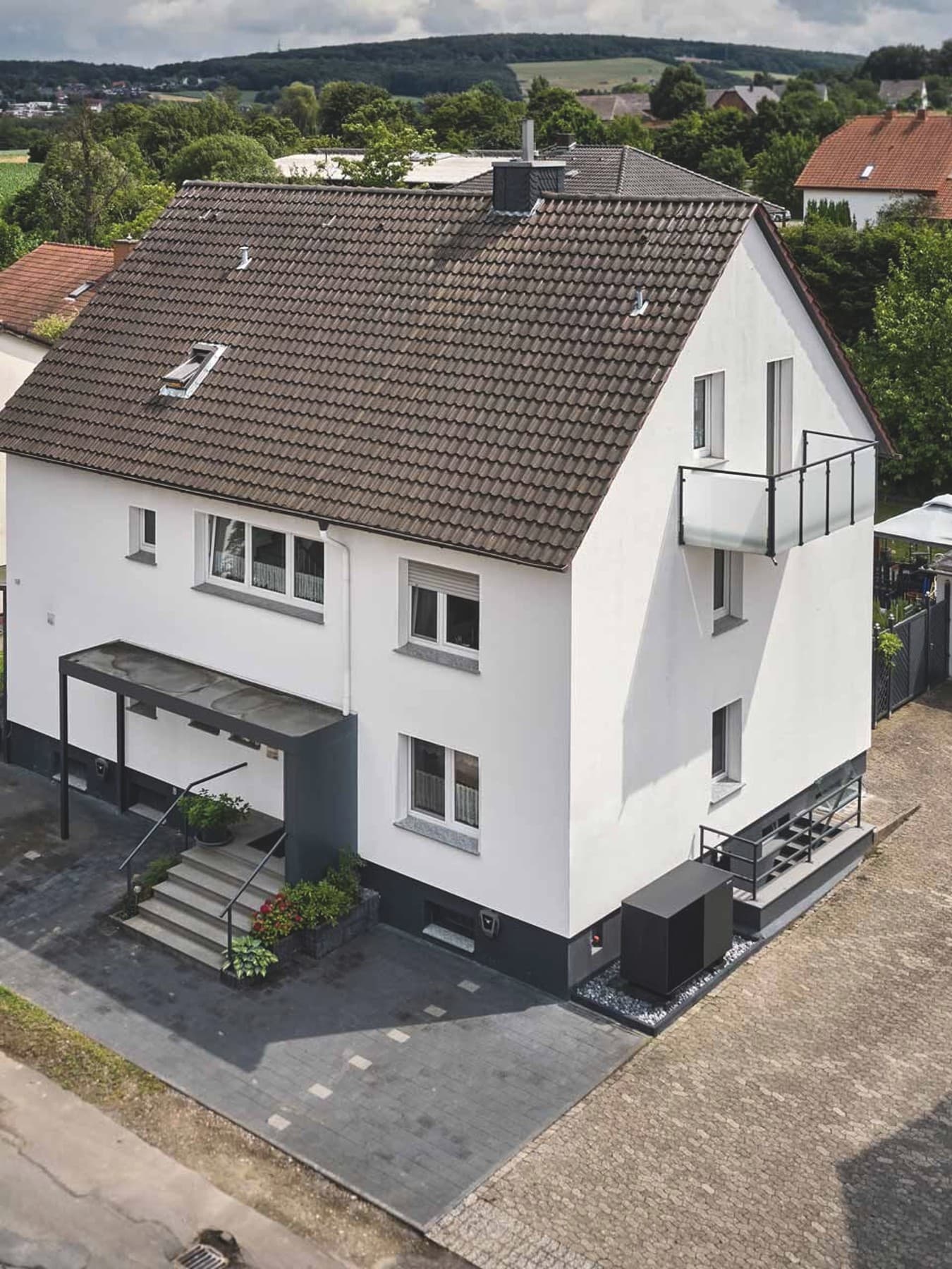Luftaufnahme eines modernisierten weißen Mehrfamilienhauses mit dunklem Ziegeldach, Balkon und überdachtem Eingangsbereich. Rechts neben dem Gebäude ist die Außeneinheit der Wärmepumpe aroTHERM perform von Vaillant installiert; auf einer benachbarten Garage sind Solarmodule zu sehen. Im Hintergrund liegen weitere Wohnhäuser, Gärten und eine grüne Landschaft.
