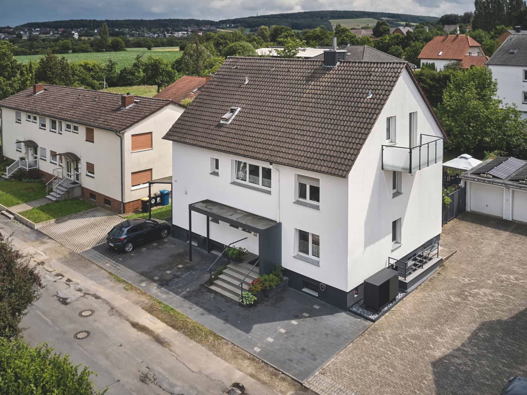 Luftaufnahme eines modernisierten weißen Mehrfamilienhauses mit dunklem Ziegeldach, Balkon und überdachtem Eingangsbereich. Rechts neben dem Gebäude ist die Außeneinheit der Wärmepumpe aroTHERM perform von Vaillant installiert; auf einer benachbarten Garage sind Solarmodule zu sehen. Im Hintergrund liegen weitere Wohnhäuser, Gärten und eine grüne Landschaft.