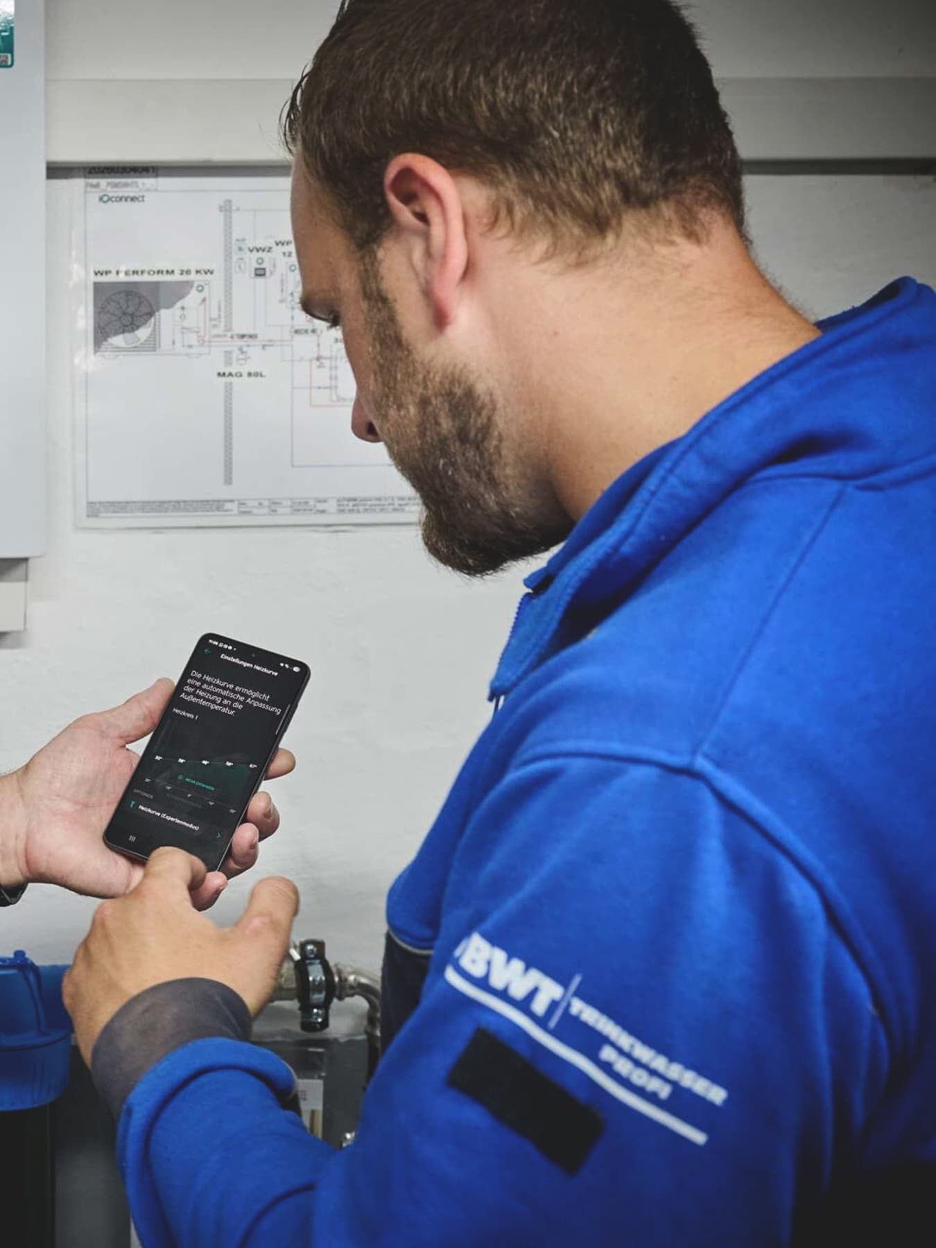 Ein Hauseigentümer und ein Fachhandwerker stehen in einem Technikraum vor der Heizungssteuerung. Der Eigentümer hält ein Smartphone mit geöffneter Steuerungs-App in der Hand, während der Fachhandwerker auf das Display zeigt und die Bedienung erklärt. Im Hintergrund sind das digitale Bedienfeld der Heizungsanlage, ein technischer Schaltplan und Teile der Installation zu sehen.