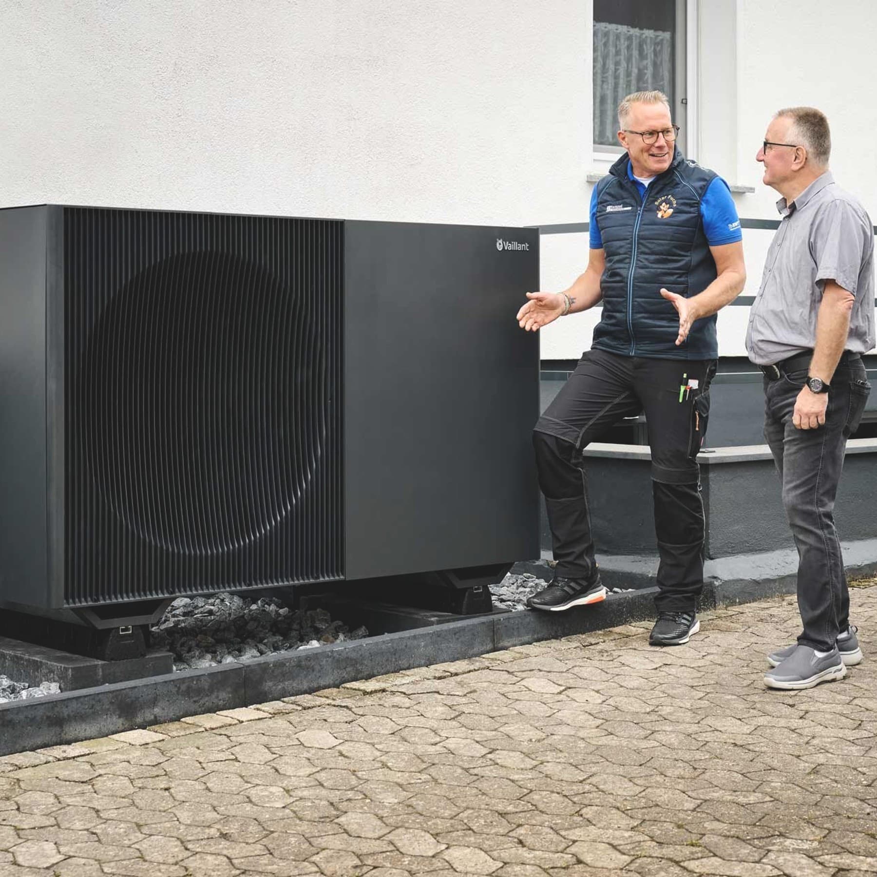 Drei Männer stehen vor der großen schwarzen Außeneinheit der aroTHERM perform von Vaillant an einem Wohnhaus. Ein Fachhandwerker in Arbeitskleidung erklärt die Anlage mit einer Handbewegung, während Eigentümer und ein Verkaufsberater von Vaillant aufmerksam zuhören. Die Wärmepumpe ist auf einem Kiesbett direkt an der Hauswand installiert; rechts ist teilweise ein weißes Fahrzeug zu sehen.