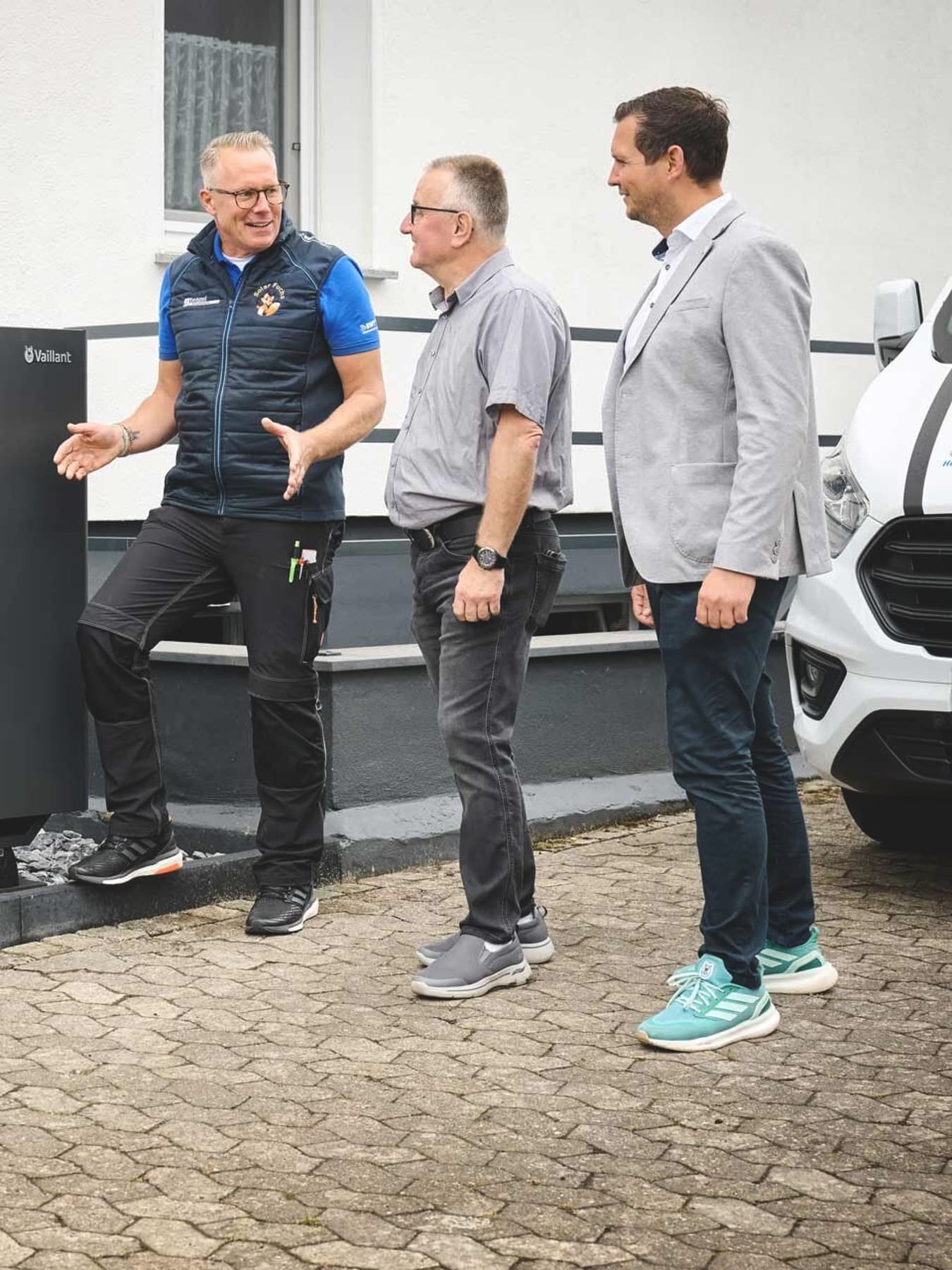 Drei Männer stehen vor der großen schwarzen Außeneinheit der aroTHERM perform von Vaillant an einem Wohnhaus. Ein Fachhandwerker in Arbeitskleidung erklärt die Anlage mit einer Handbewegung, während Eigentümer und ein Verkaufsberater von Vaillant aufmerksam zuhören. Die Wärmepumpe ist auf einem Kiesbett direkt an der Hauswand installiert; rechts ist teilweise ein weißes Fahrzeug zu sehen.