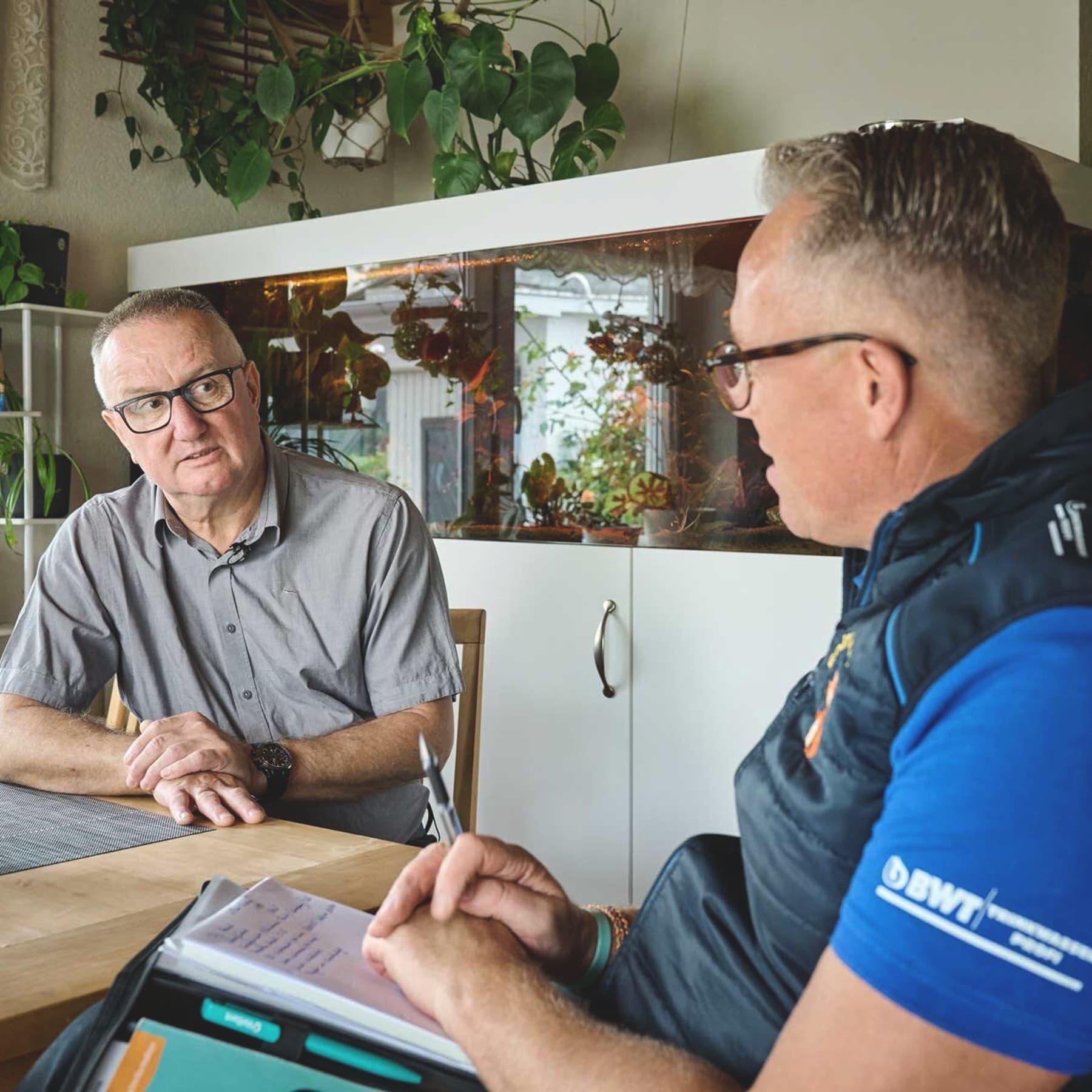 Ein Hauseigentümer und ein Fachhandwerker sitzen sich an einem Holztisch gegenüber und führen ein Beratungsgespräch. Der Eigentümer links trägt ein graues Hemd und eine Brille und hört mit gefalteten Händen aufmerksam zu. Der Fachhandwerker rechts trägt ein blaues Shirt mit dunkler Weste und hält einen Stift über seinen Notizen. Im Hintergrund stehen ein großes Aquarium sowie zahlreiche grüne Zimmerpflanzen.