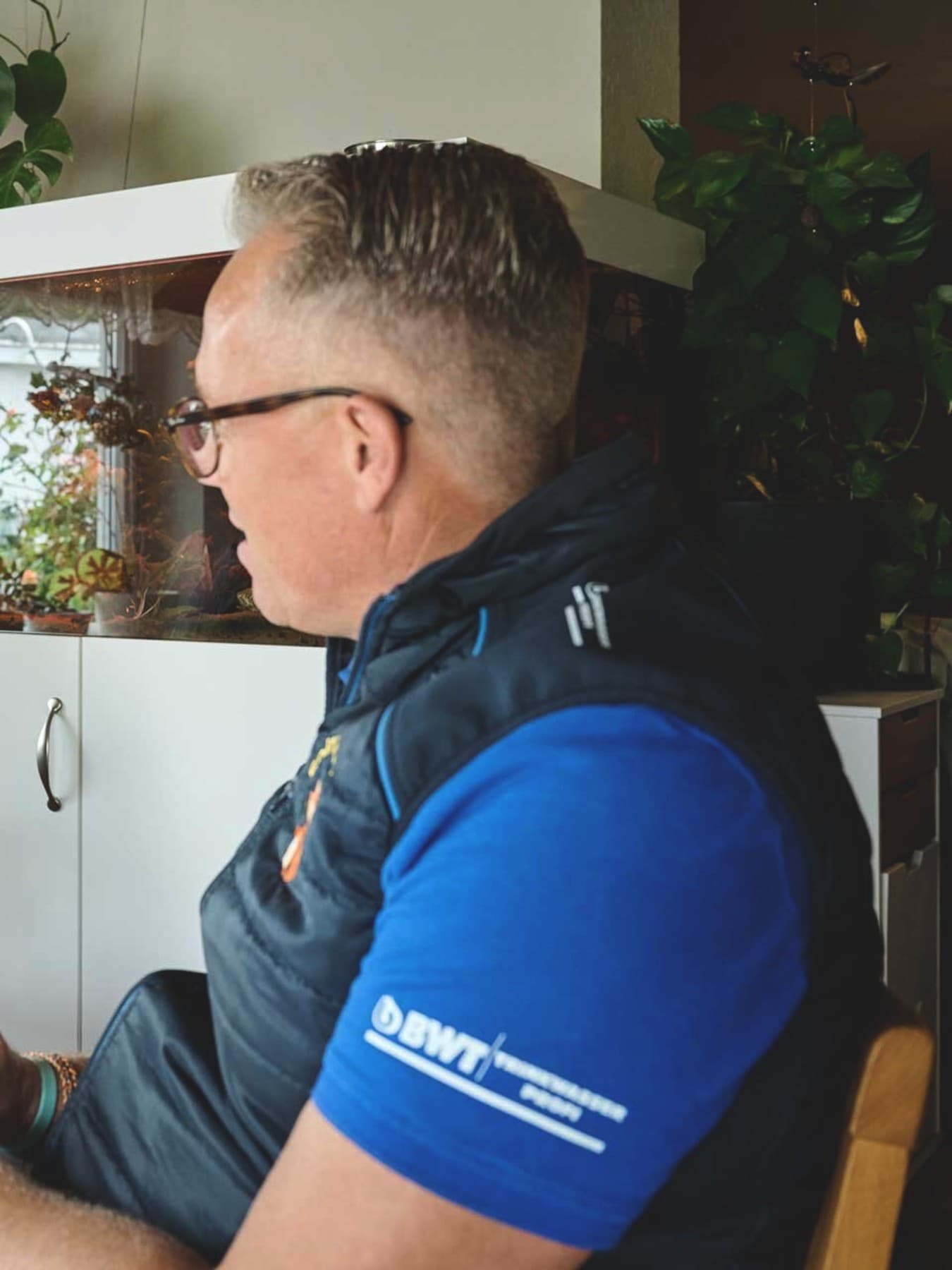 Ein Hauseigentümer und ein Fachhandwerker sitzen sich an einem Holztisch gegenüber und führen ein Beratungsgespräch. Der Eigentümer links trägt ein graues Hemd und eine Brille und hört mit gefalteten Händen aufmerksam zu. Der Fachhandwerker rechts trägt ein blaues Shirt mit dunkler Weste und hält einen Stift über seinen Notizen. Im Hintergrund stehen ein großes Aquarium sowie zahlreiche grüne Zimmerpflanzen.