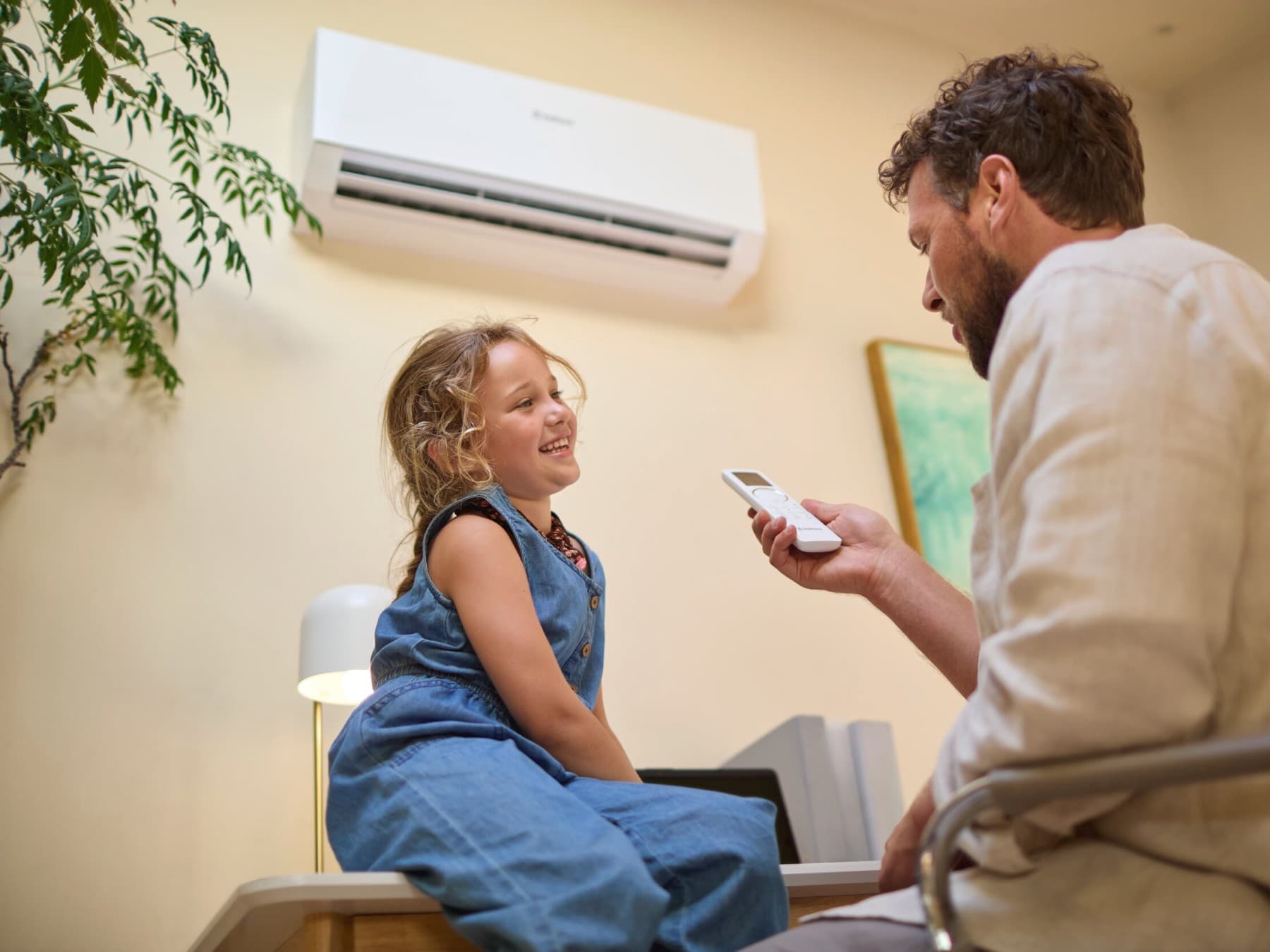 Ein Mann hält eine Fernbedienung in der Hand. Ein Mädchen schaut ihm dabei zu, wie er damit die Vaillant Klimaanlage climaVAIR plus bedient, die im Hintergrund an der Wand hängt.