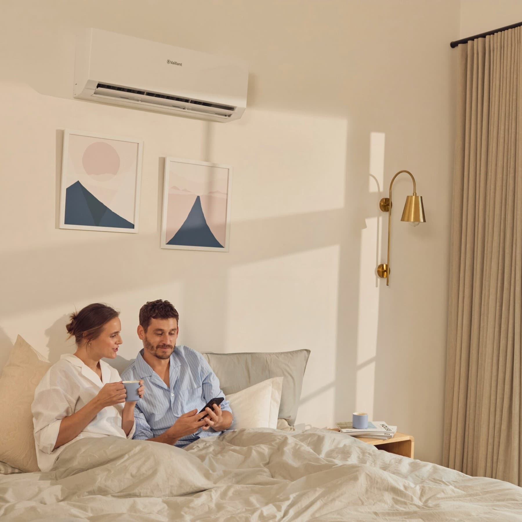 Paar sitzt im Schlafzimmer. Über dem Bett hängt die Innenheit der Klimaanlage climaVAIR plus von Vaillant und vor dem Fenster ist die Außeneinheit zu sehen.