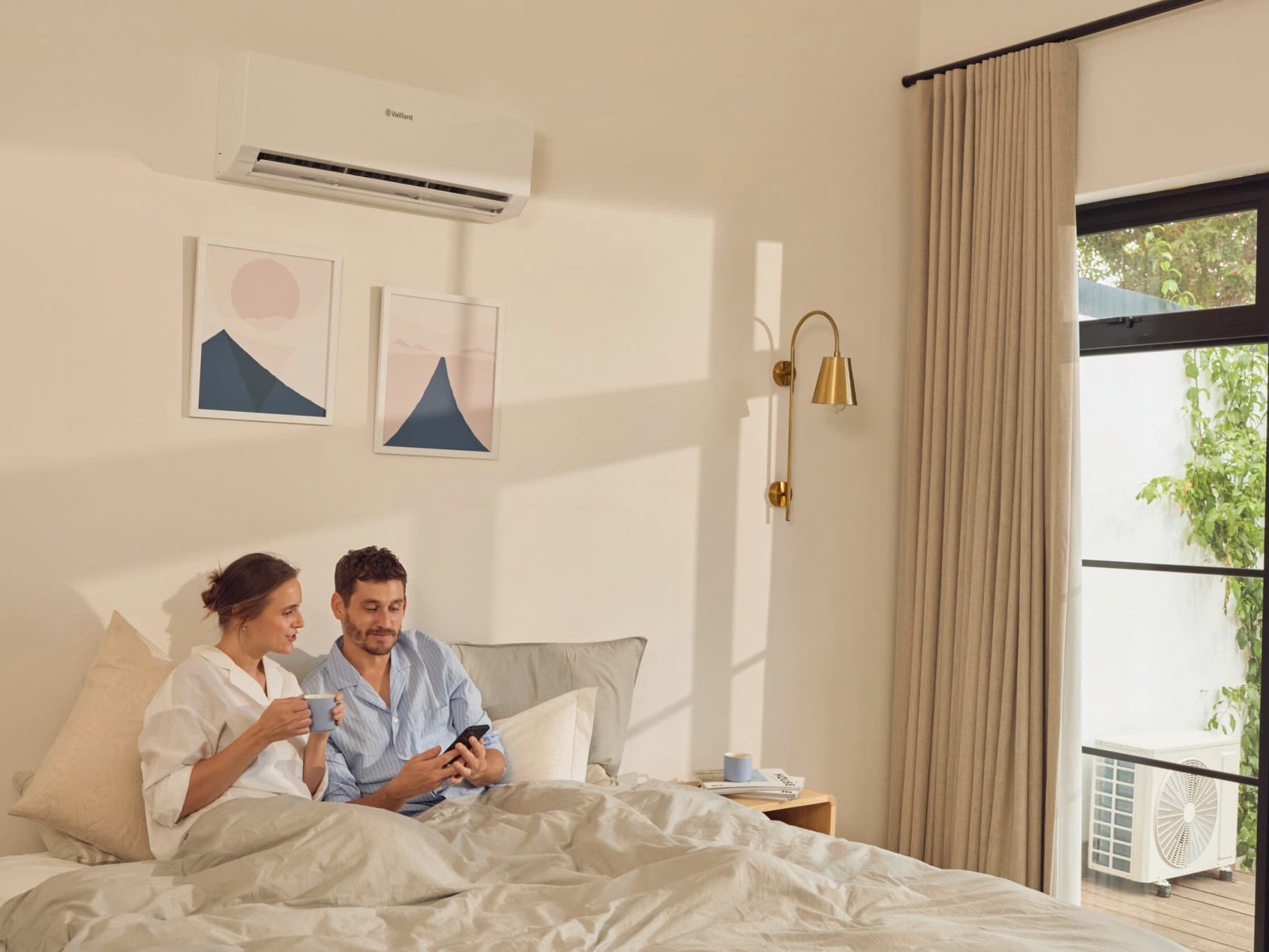 Paar sitzt im Schlafzimmer. Über dem Bett hängt die Innenheit der Klimaanlage climaVAIR plus von Vaillant und vor dem Fenster ist die Außeneinheit zu sehen.