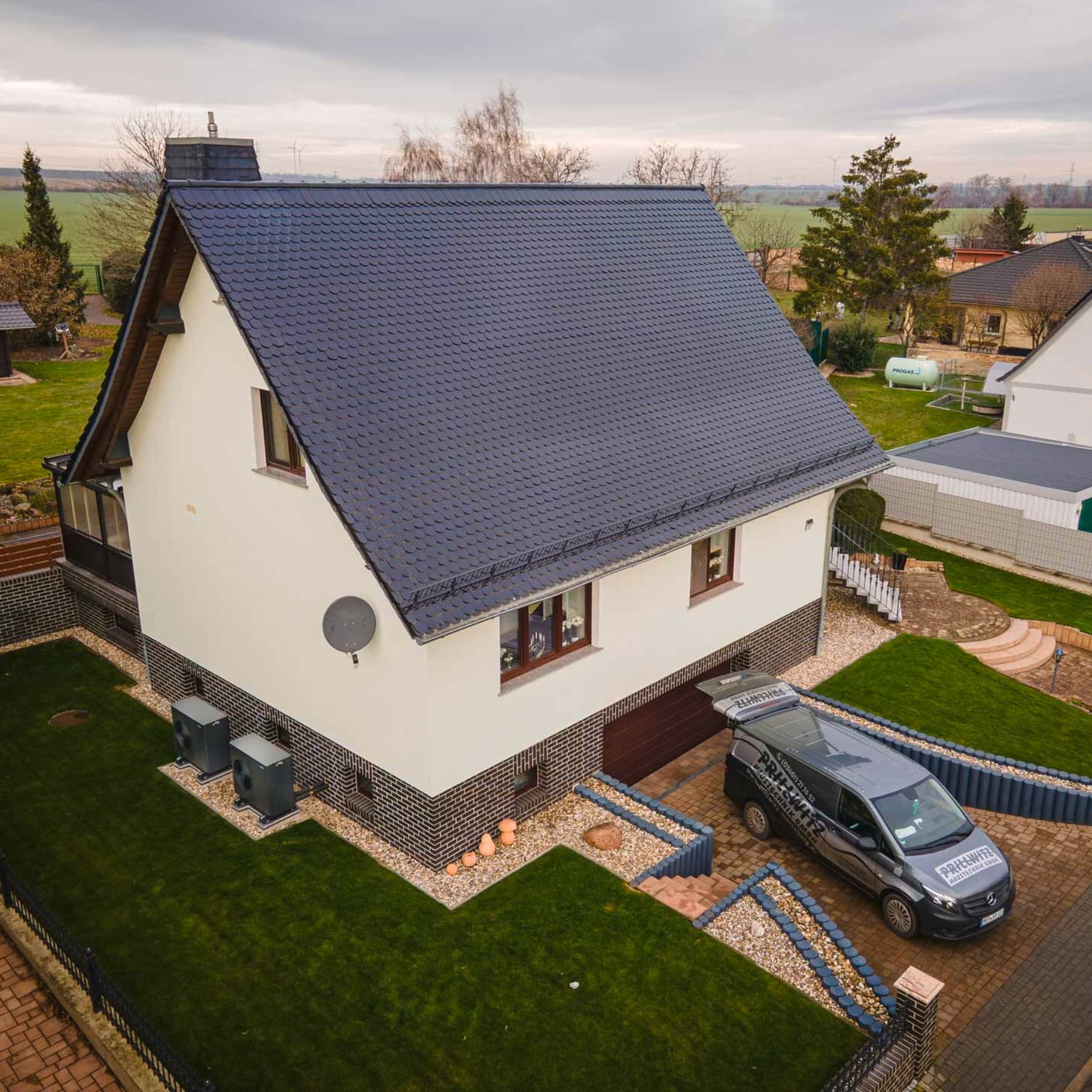 Freistehendes Einfamilienhaus mit hellem Fassadenanstrich und dunklem Dach, umgeben von einem gepflegten Garten mit Rasenflächen. Vor dem Haus befindet sich eine gepflasterte Einfahrt mit geparktem Fahrzeug, im Hintergrund sind weitere Wohnhäuser und eine ländliche Umgebung zu sehen