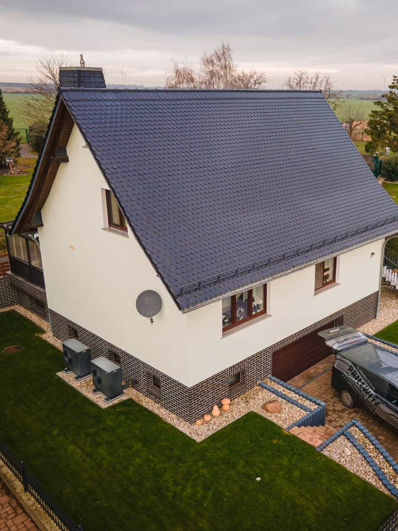 Freistehendes Einfamilienhaus mit hellem Fassadenanstrich und dunklem Dach, umgeben von einem gepflegten Garten mit Rasenflächen. Vor dem Haus befindet sich eine gepflasterte Einfahrt mit geparktem Fahrzeug, im Hintergrund sind weitere Wohnhäuser und eine ländliche Umgebung zu sehen