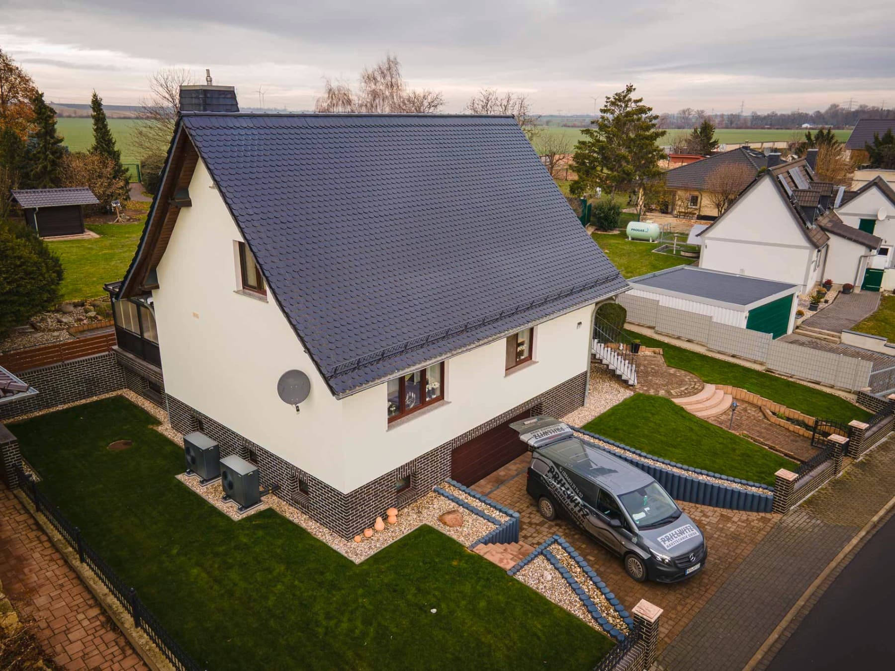 Freistehendes Einfamilienhaus mit hellem Fassadenanstrich und dunklem Dach, umgeben von einem gepflegten Garten mit Rasenflächen. Vor dem Haus befindet sich eine gepflasterte Einfahrt mit geparktem Fahrzeug, im Hintergrund sind weitere Wohnhäuser und eine ländliche Umgebung zu sehen
