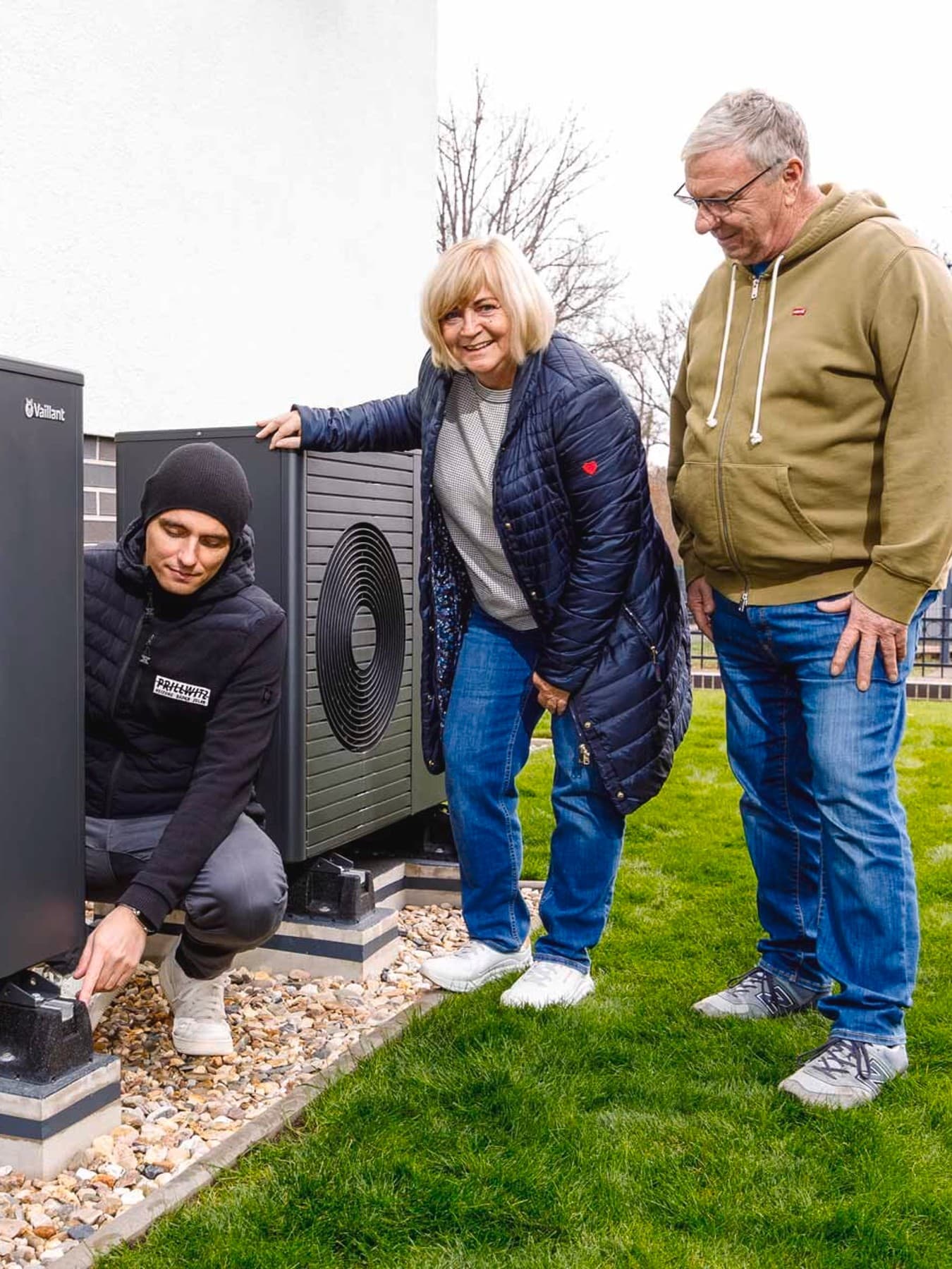 Außenszene auf einem Grundstück: Ein Monteur kniet neben einer installierten Wärmepumpe und prüft diese, während eine Frau und ein Mann danebenstehen und zusehen. Die Geräte stehen auf Fundamenten im Garten neben einem Wohnhaus, im Hintergrund sind weitere Häuser zu erkennen