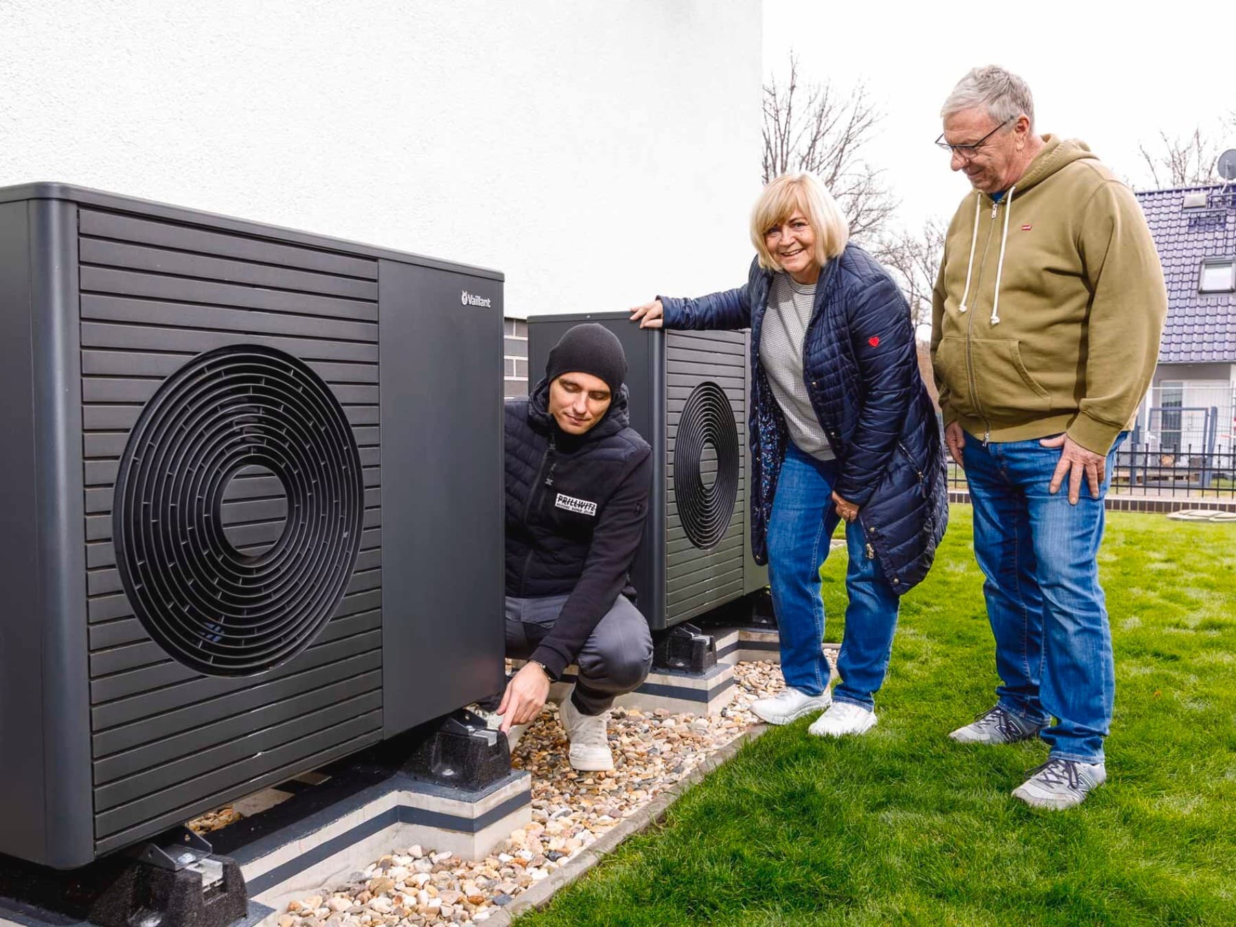 Außenszene auf einem Grundstück: Ein Monteur kniet neben einer installierten Wärmepumpe und prüft diese, während eine Frau und ein Mann danebenstehen und zusehen. Die Geräte stehen auf Fundamenten im Garten neben einem Wohnhaus, im Hintergrund sind weitere Häuser zu erkennen