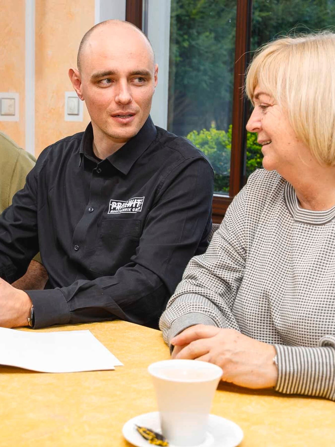 Drei Personen sitzen an einem Tisch in einem hellen Wohnraum. Ein Mann mit schwarzem Hemd hat ein Tablet in der Hand und berät eine Frau und ihren Mann, die jeweils rechts und links von ihm sitzen. Sie trägt einen grauen Pulli und einen blonden Bob, ihr Mann wiederum einen grünen Pulli sowie graue kurze Haare. Auf dem Tisch liegen Dokumente, daneben stehen Kaffeetassen und Gebäck.