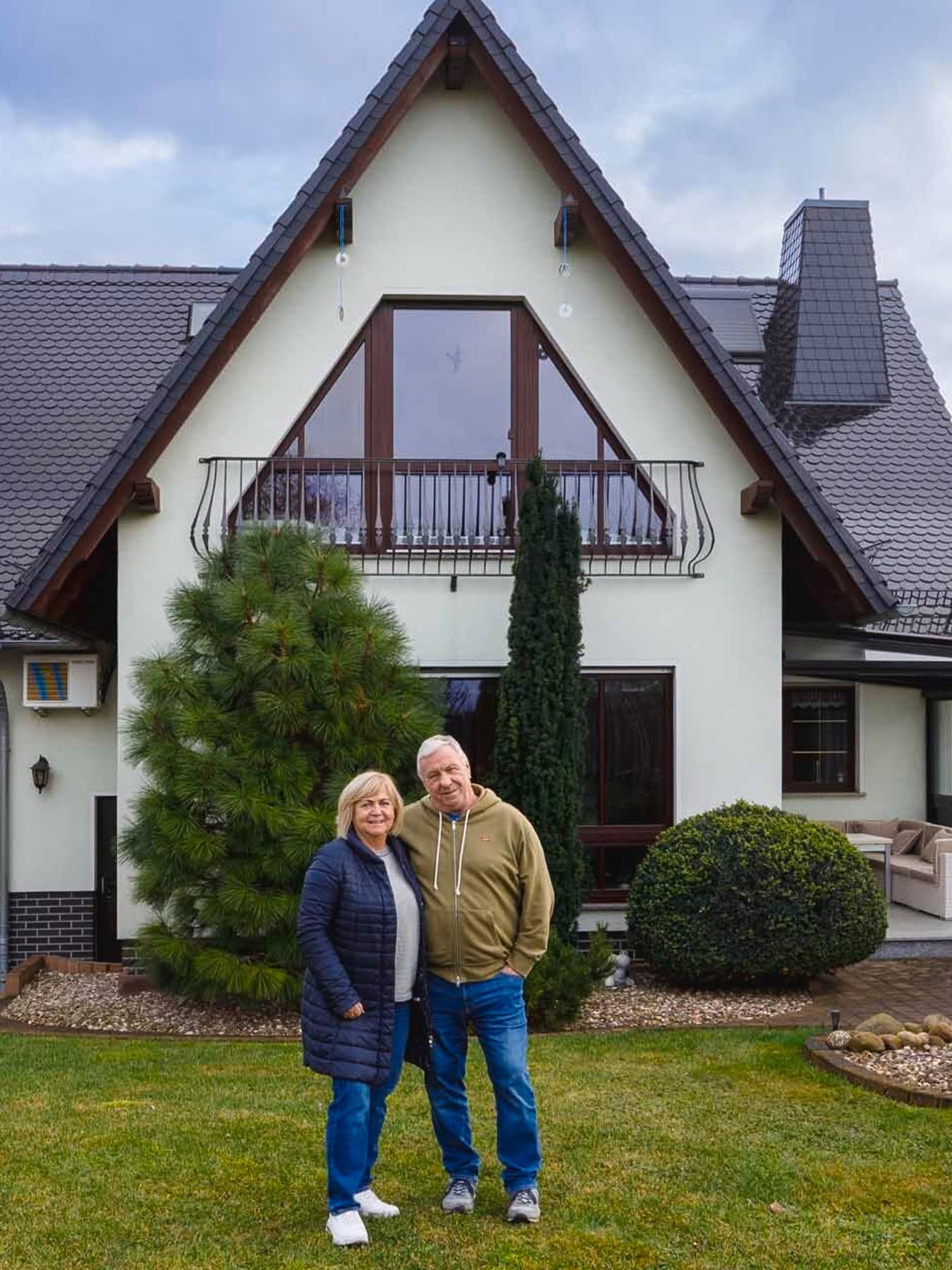 Ein älteres Paar steht auf einer Rasenfläche vor einem freistehenden Einfamilienhaus mit hellem Fassadenanstrich und dunklem Dach. Das Haus verfügt über große Fenster, einen Balkon im Obergeschoss sowie eine überdachte Terrasse. Der Garten ist gepflegt, mit Bäumen, Sträuchern und angelegten Beeten