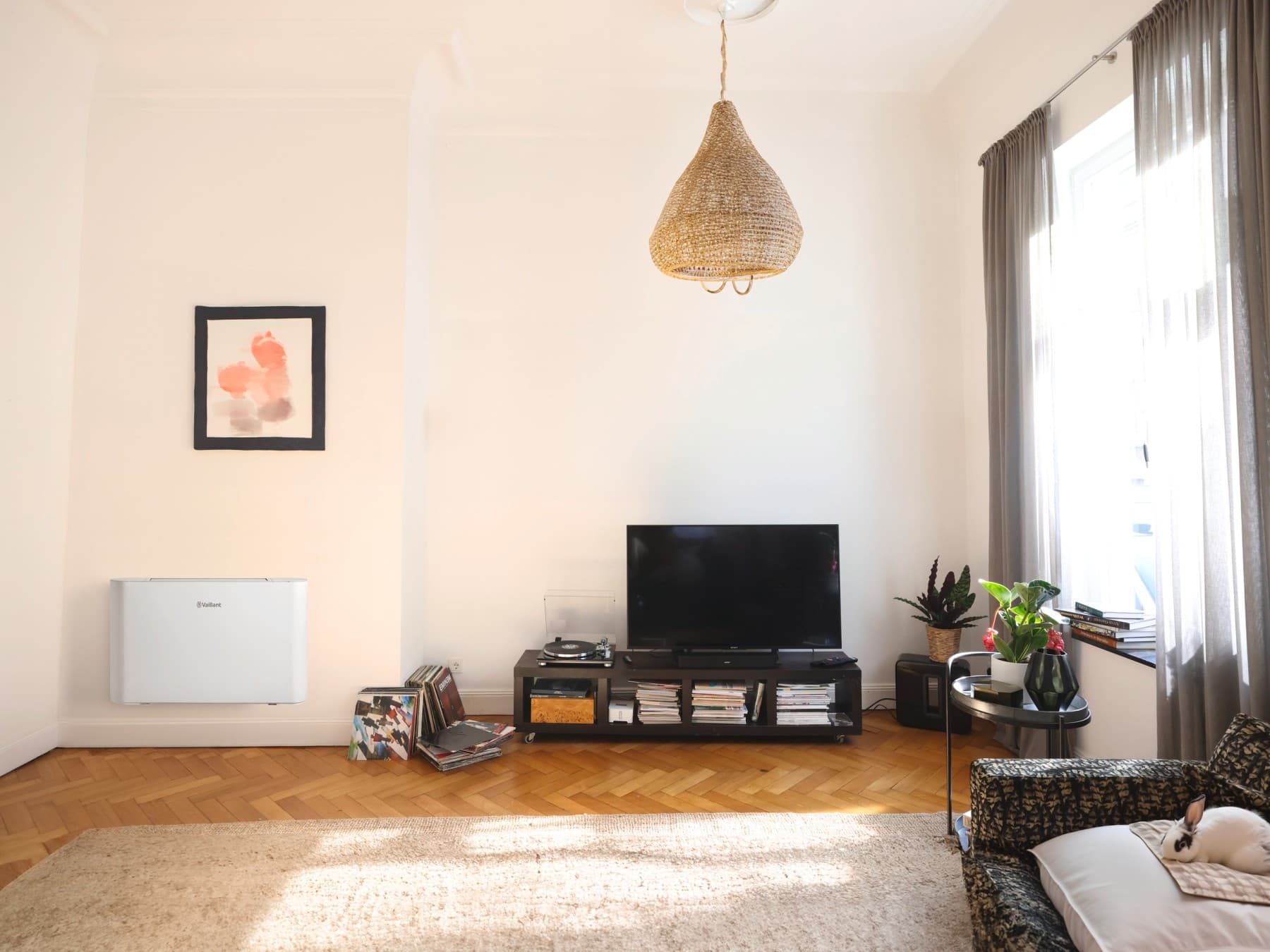 Wandmontierter Konsole-Gebläsekonvektor aroVAIR pro CN hängt an einer Wand in Bodennähe unter einem Bild. Rechts davon steht ein Sideboard mit Plattensammlung, Plattenspieler und Fernseher.