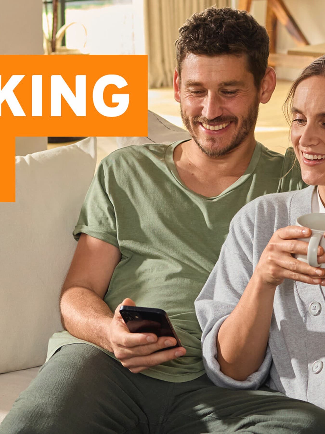 Ein Mann und eine Frau sitzen gemeinsam auf einer hellen Couch und schauen fröhlich auf ein Smartphone, das der Mann in der Hand hält. Die Frau hält eine Tasse in den Händen.