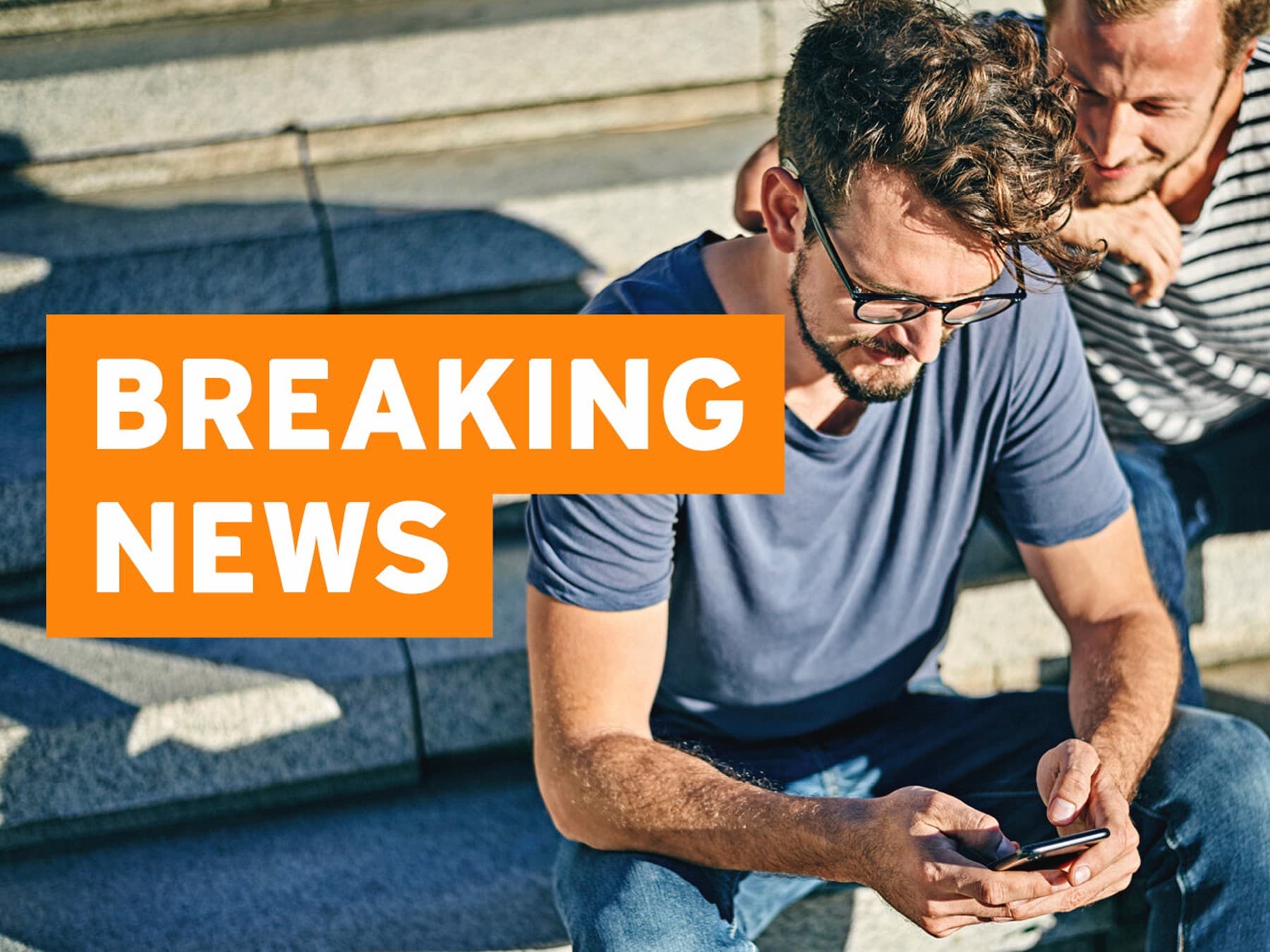 Bild zeigt zwei Männer, die auf sonnenbeschienener Treppe sitzen und gemeinsam auf ein Smartphone gucken, im Vordergrund befindet sich ein oranger Balken mit der weißen Aufschrift: breaking news