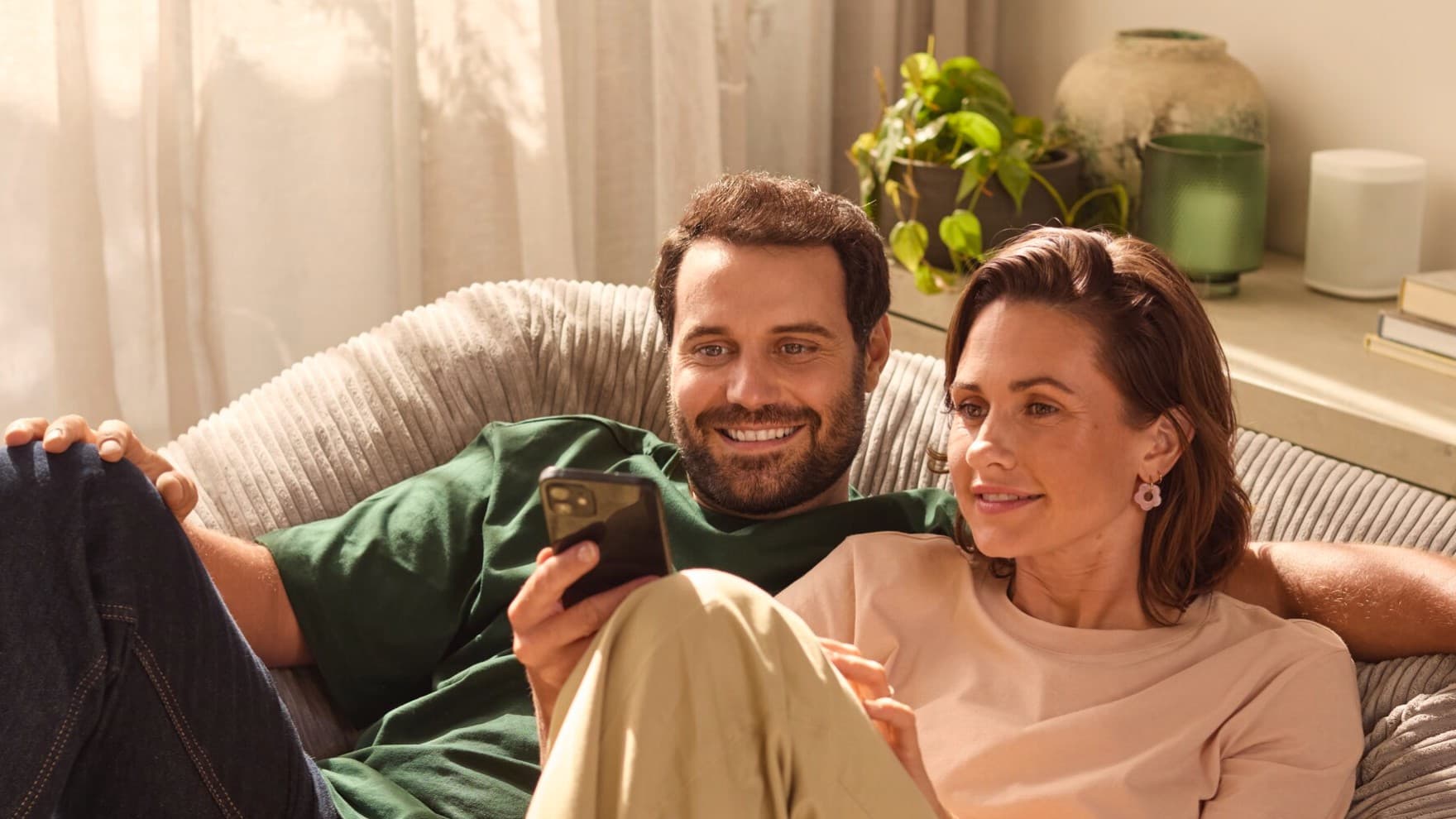 Eine Frau und ein Mann sitzen gemeinsam auf einer Couch und schauen auf ein Smartphone. Der Mann hat der Frau den Arm um die Schulter gelegt und lächelt. Im Hintergrund sieht man eine grüne Pflanze und farblich passende Deko auf einem Sideboard sowie eine weiße Gardine.