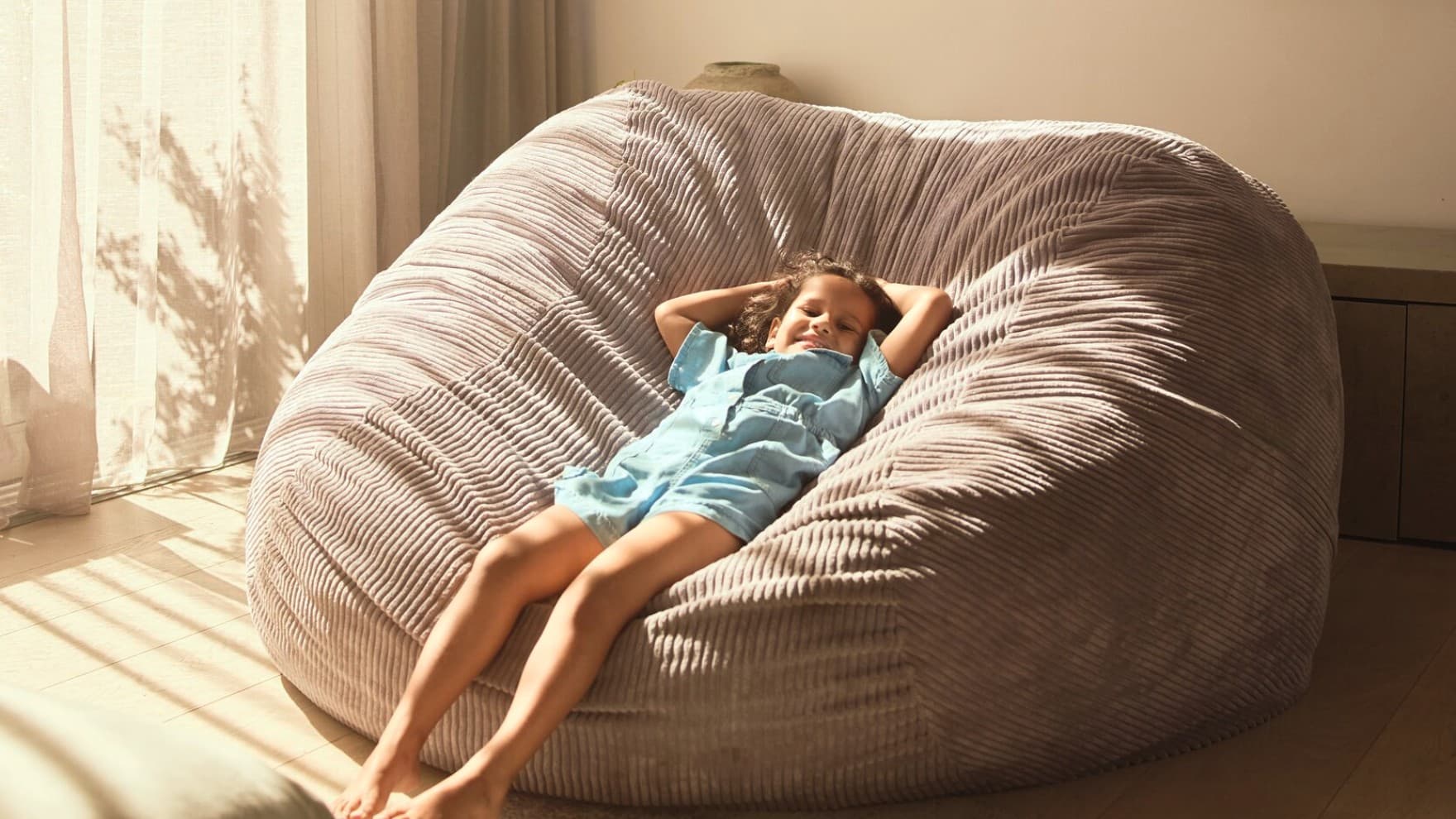 Ein lächelndes Kind liegt in einem großen Sitzsack aus Cord, der in der Ecke in einem Zimmer vor einem Fenster mit bodenlangen Gardinen steht.