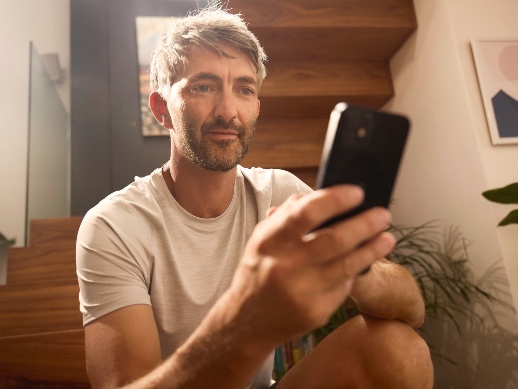 Ein Mann sitzt auf einer Holztreppe in einem Haus und schaut auf sein Smartphone.