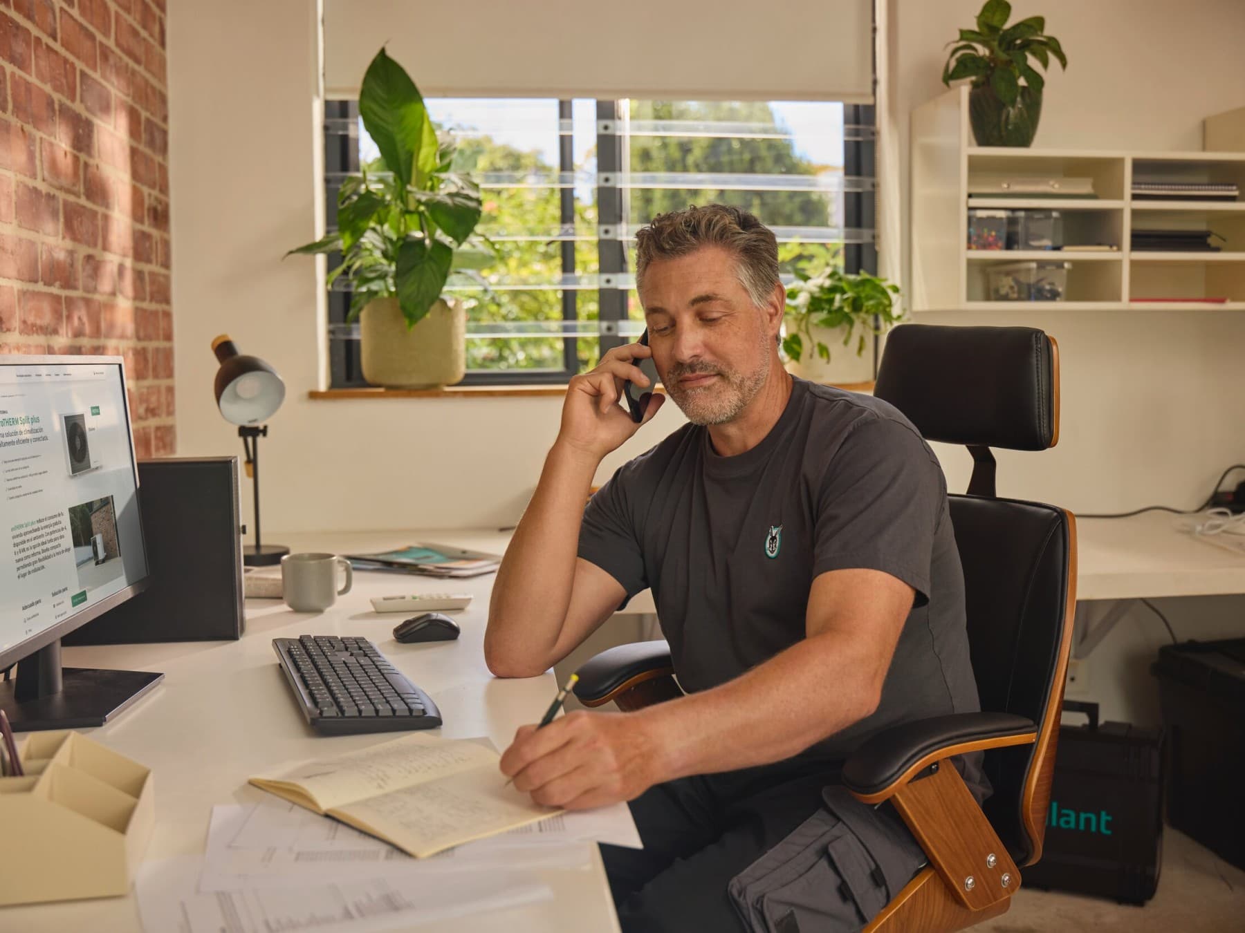 Ein Mann mit Vaillant T-Shirt sitzt telefonierend an einem Schreibtisch vor einem Computerbildschirm und macht sich Notizen in ein Notizbuch.