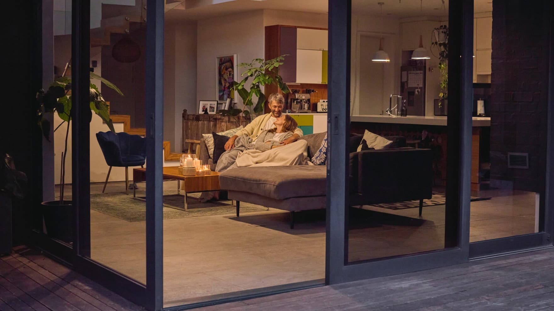 Aufnahme von draußen ins Haus. Ein Ehepaar sitzt auf der Couch bei weit geöffneter Türe zum Lüften.