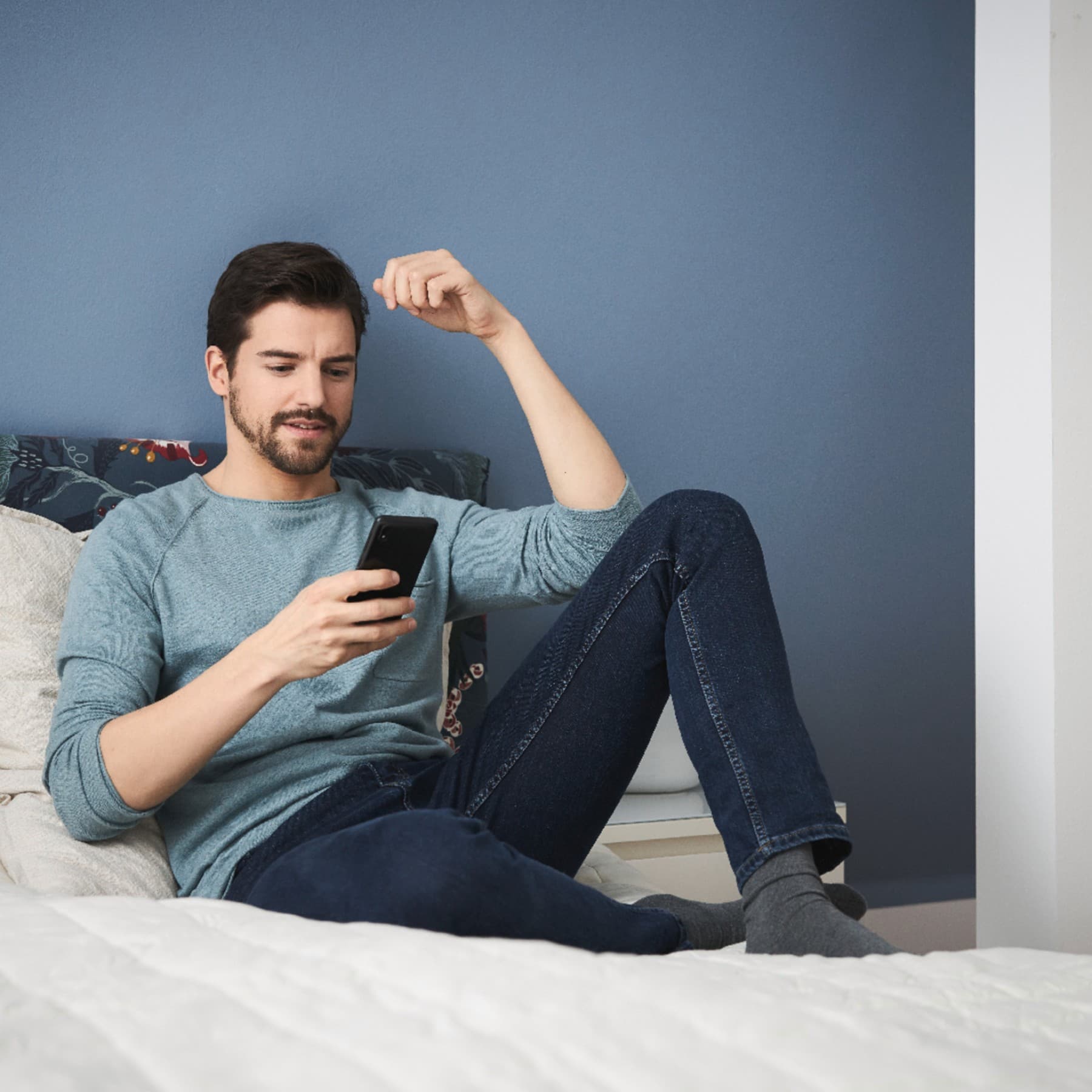 Mann sitzt mit Handy in der Hand auf einem Bett
