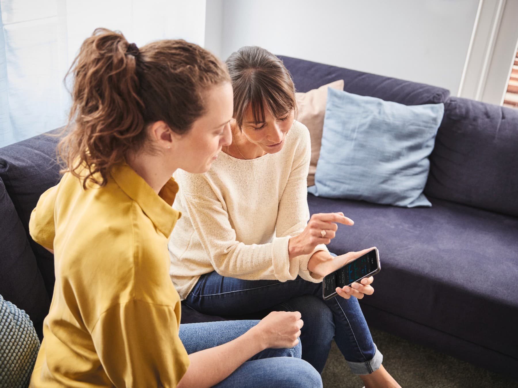 Zwei Frauen sitzen auf einem Sofa in einem Wohnzimmer. Die eine hält ein Smartphone in der Hand und zeigt der anderen die myVAILLANT App.