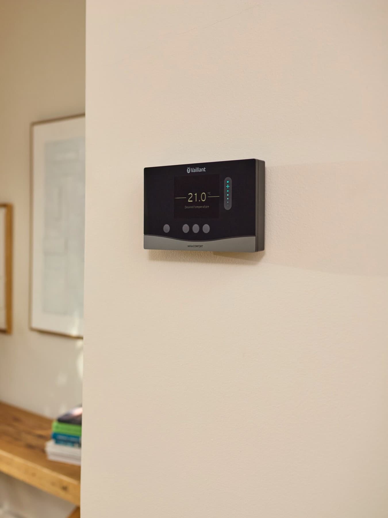 Der Vaillant Regler sensoCOMFORT VRC 720 hängt an einer weißen Wand. Im Hintergrund erkennt man eine Wohnzimmer-Ecke mit einem Holztisch, auf dem eine Pflanze steht. An der Wand hängen Bilderrahmen. Links im Bild sieht man ein bodenhohes Fenster mit dem Blick in einen grünen Garten. Am Fenster, hinter der kurzen Tischkante, hängt ein bodenlanger Vorhang.