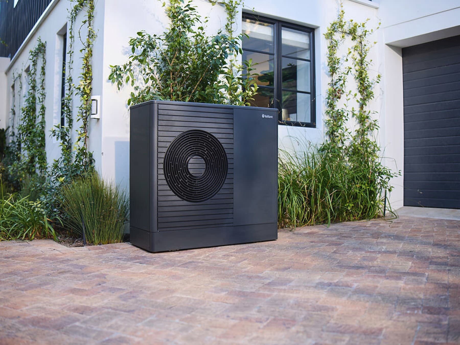 Die Inverter-Wärmepumpe aroTHERM plus von Vaillant auf der Garagenauffahrt eines weißen Hauses.