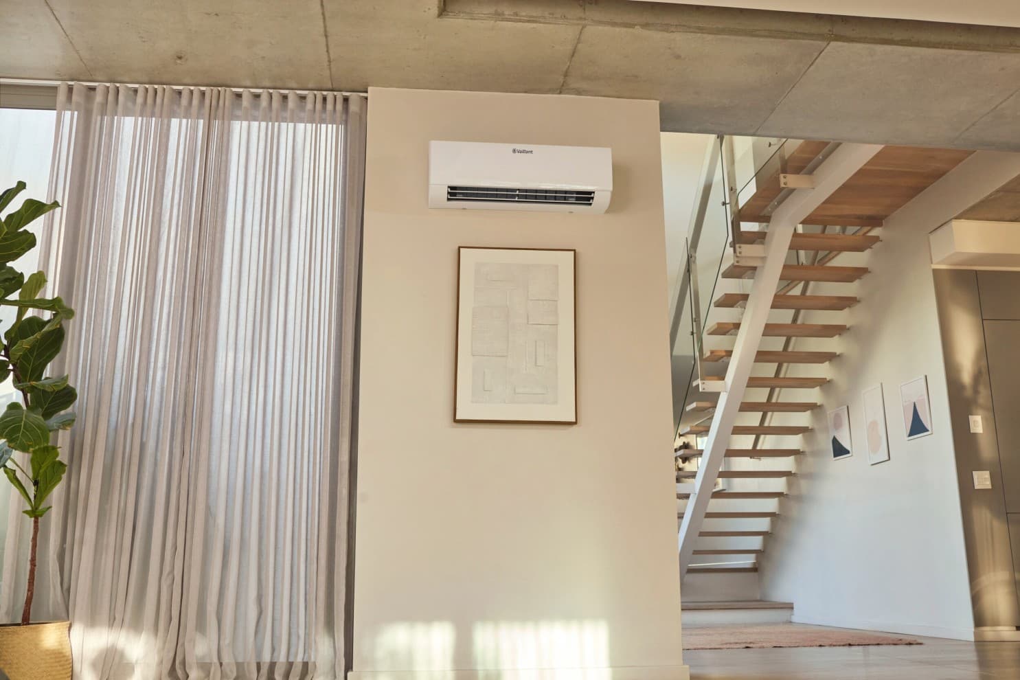Wand-Gebläsekonvektor aroVAIR pro WN hängt an einer Wand hoch oben unter der Decke über einem Bild neben einem Fenster, das mit einem Gardinenvorhang zugezogen ist. Rechts im Bild ist ein Treppenaufgang des Hauses zu sehen.