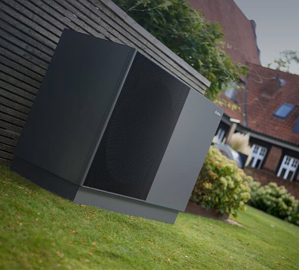 Die neue Vaillant Wärmepumpe aroTHERM perform steht auf einem gepflegten rasen vor einem Gartenzaun. Im Hintergrund sind Blumenbeete und ein Haus zu sehen.
