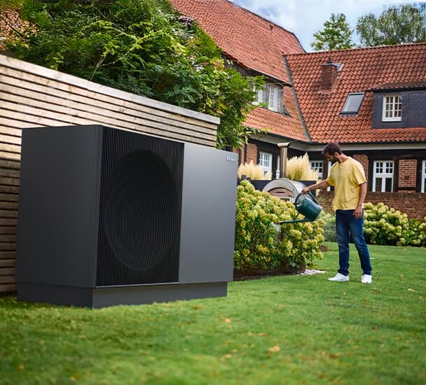 Eine Vaillant Wärmepumpe aroTHERM perform steht auf einem gepflegten Rasen vor einem Gartenzaun neben Blumenbeeten. Ein Mann steht vor dem Beet und gießt die Blumen mit einer Gießkanne.