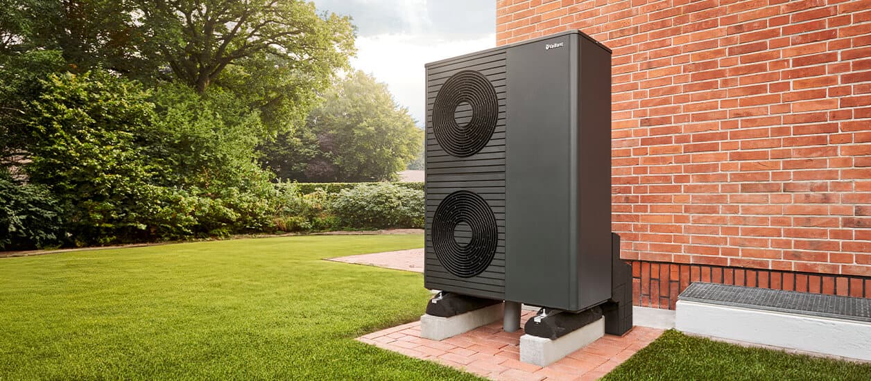 Die neue Vaillant Wärmepumpe aroTHERM plus vor einer Backsteinwand in einem gepflegten Garten, links sind Büsche und Bäume sowie ein gemähter Rasen zu sehen.