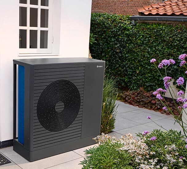 Die neue Vaillant Wärmepumpe aroTHERM pro steht unter einem Fenster vor einer weißen Hauswand zwischen Blumenbeeten.