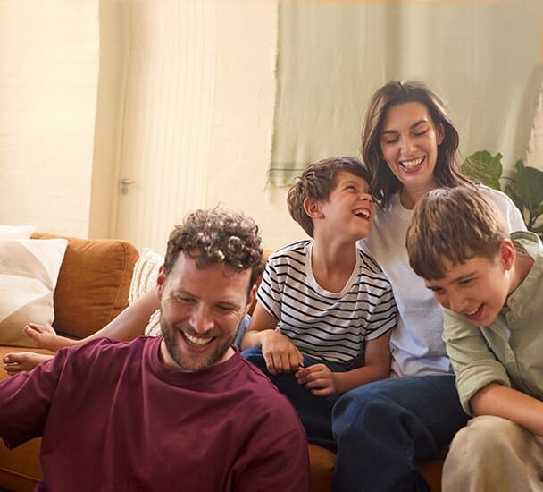 Eine Familie, bestehend aus zwei Jungs, einer Frau und einem Mann, sitzen in einem Wohnzimmer vor und auf einem Sofa. Im Hintergrund ist ein Fenster mit Gardine und Pflanzen zu sehen.