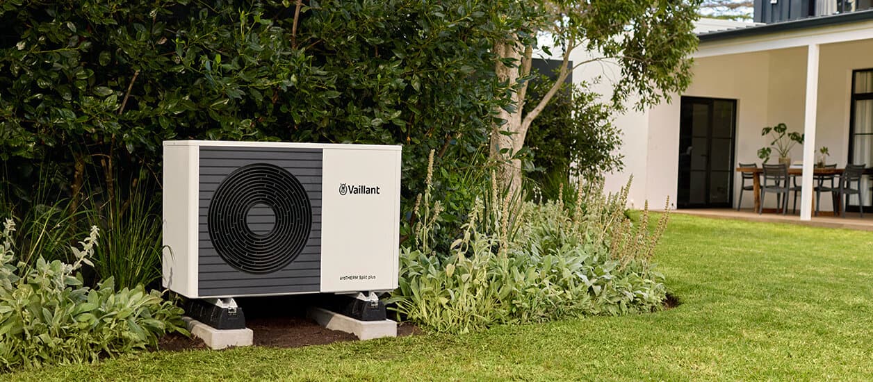 Die Vaillant Wärmepumpe aroTHERM split plus steht in einem grünen Garten neben Büschen, rechts im Bild ist ein Teil eines modernen Hauses zu sehen.