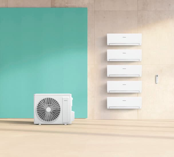 Eine climaVAIR Außenheinheit steht vor grünem Hintergrund. Daneben hängen an einer grauen Wand mehrere Wandkonsolen und eine Fernbedienung.