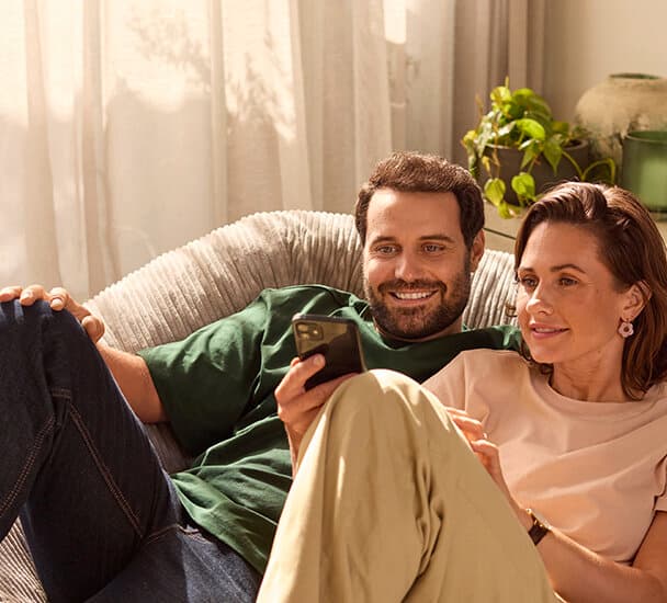 Ein Mann und eine Frau sitzen gemeinsam, Arm in Arm auf einer Couch und schauen auf ein Smartphone, das die Frau in der Hand hält.